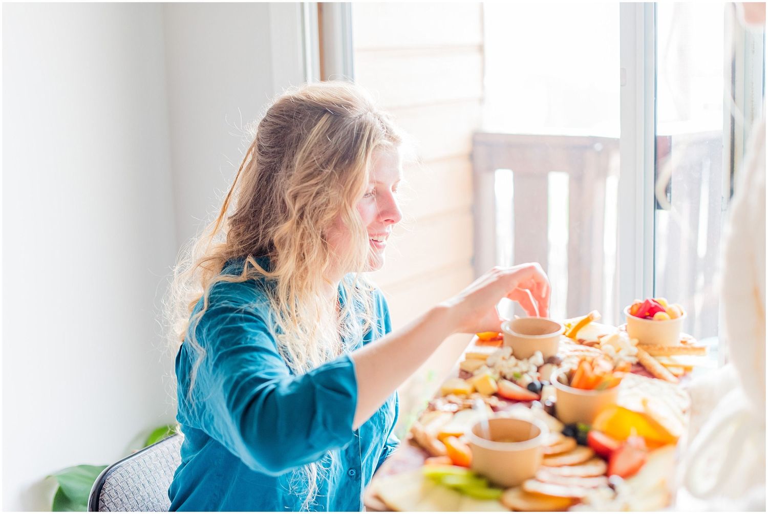 Smiling bridesmaid in teal robe reaches for food at sunlit bridal party charcuterie spread.
