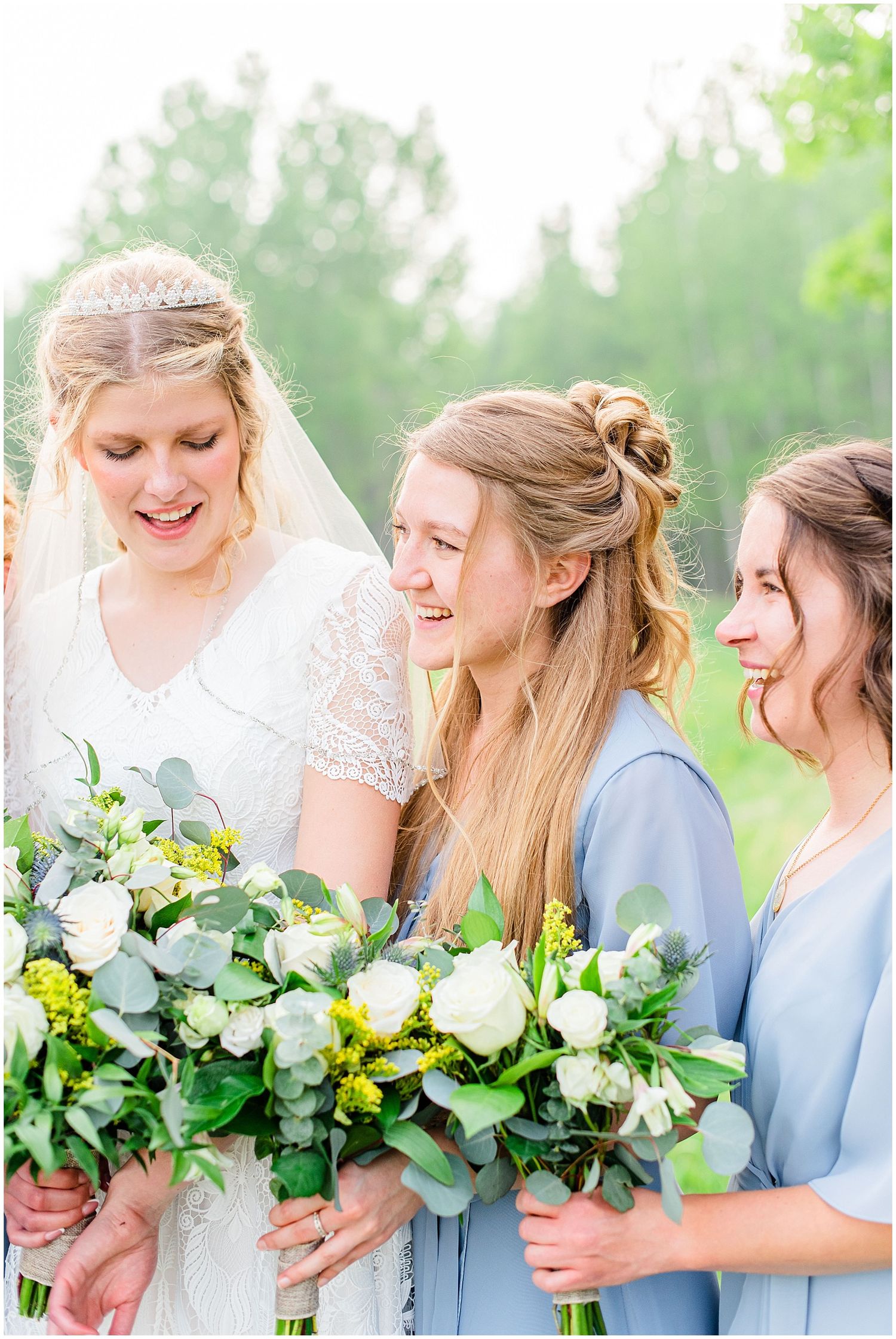 Bride in lace gown and veil laughs with bridesmaids in blue dresses holding white rose and greenery bouquets outdoors.