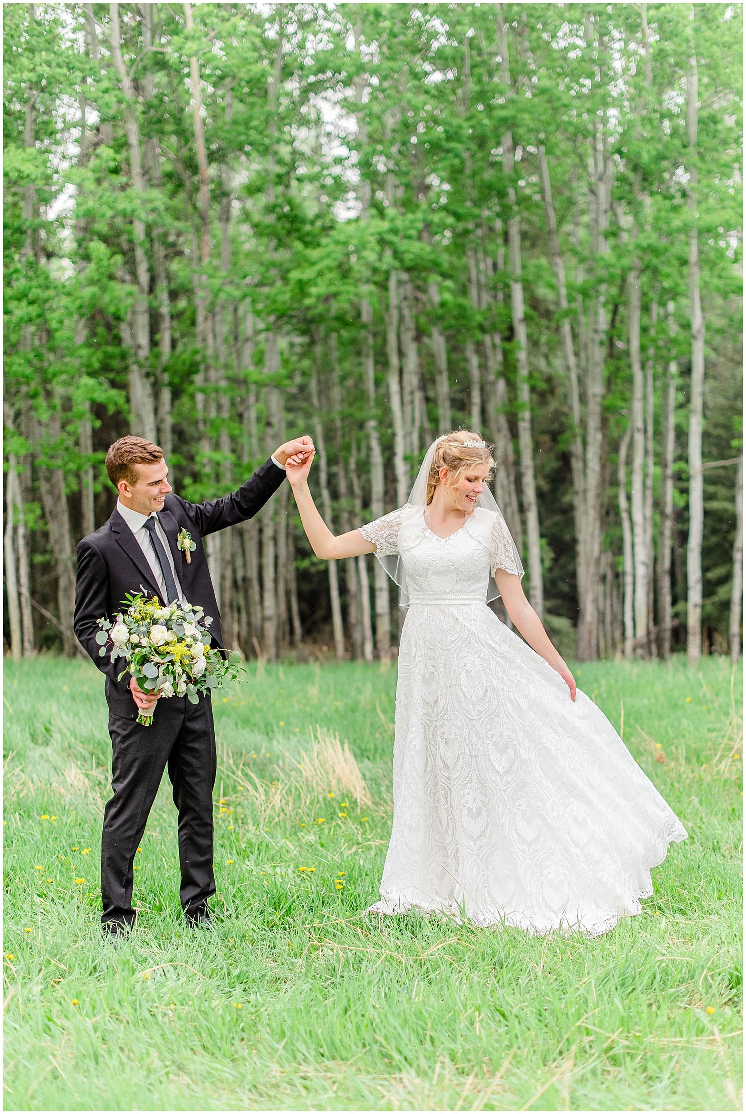 Groom in black suit twirls bride in white lace gown in a lush green meadow with birch trees.
