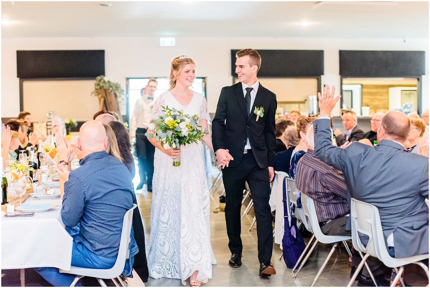 Newlyweds enter wedding reception hall hand in hand as guests applaud and celebrate.