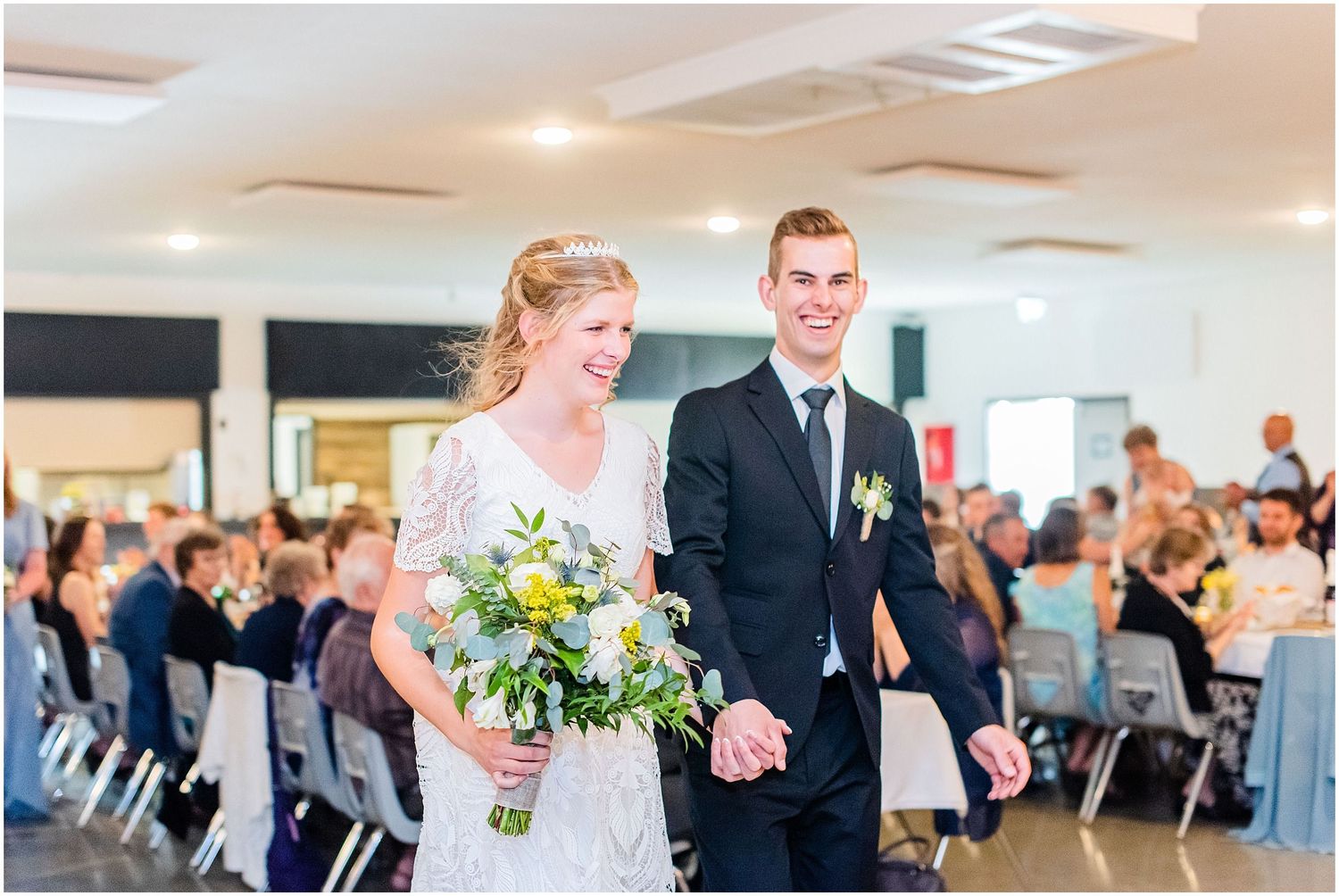 Smiling newlyweds walk hand in hand down the aisle at their wedding reception, bride holding yellow and white bouquet.