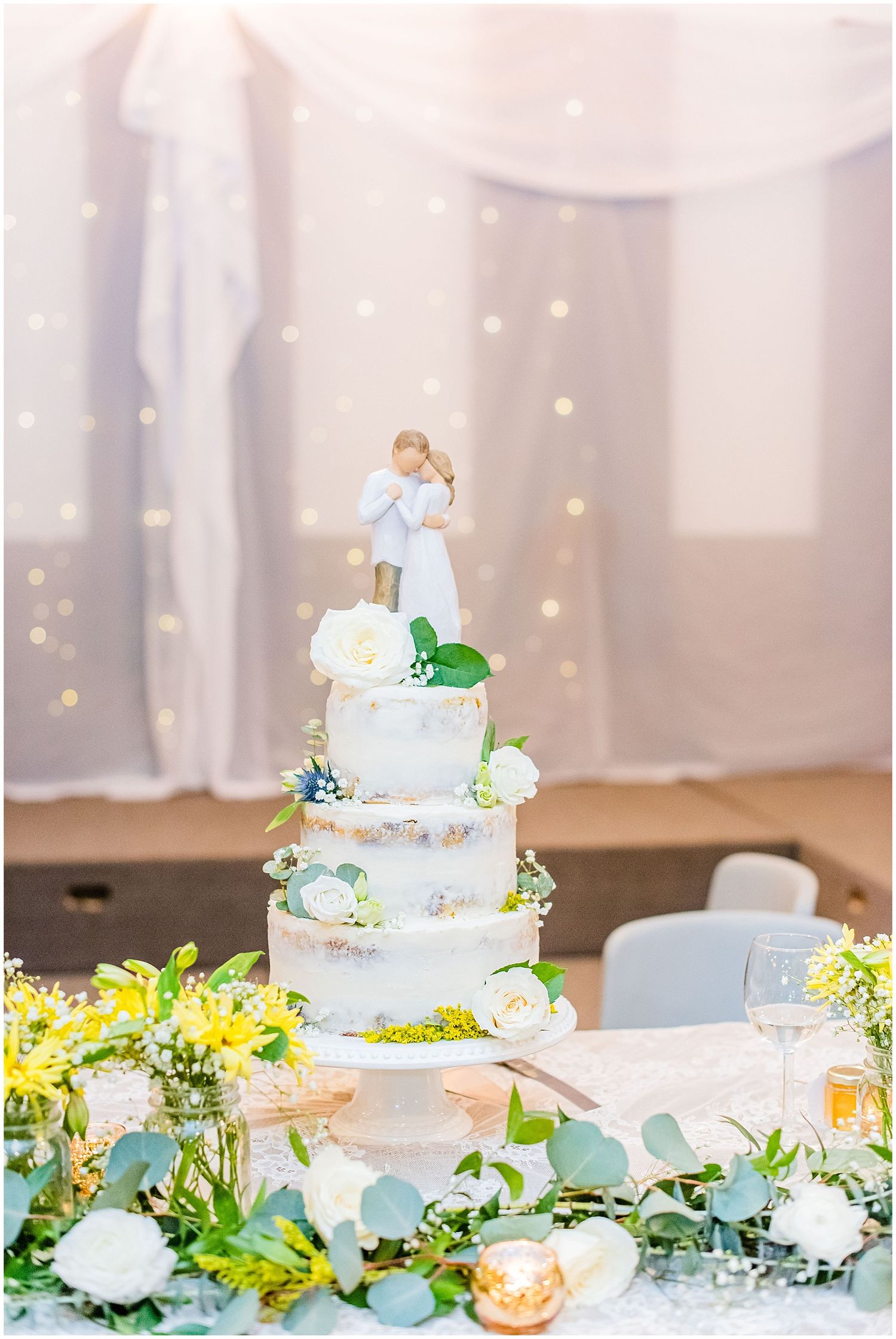Elegant four-tier white wedding cake adorned with fresh flowers and a couple figurine topper surrounded by greenery.