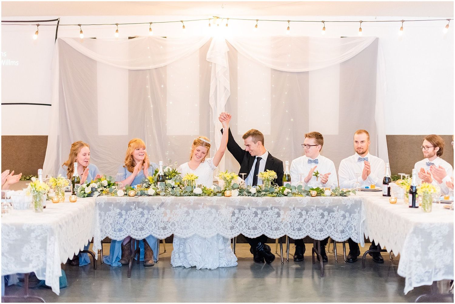 Wedding party raises glasses at elegant reception table with lace tablecloth and floral centerpieces.