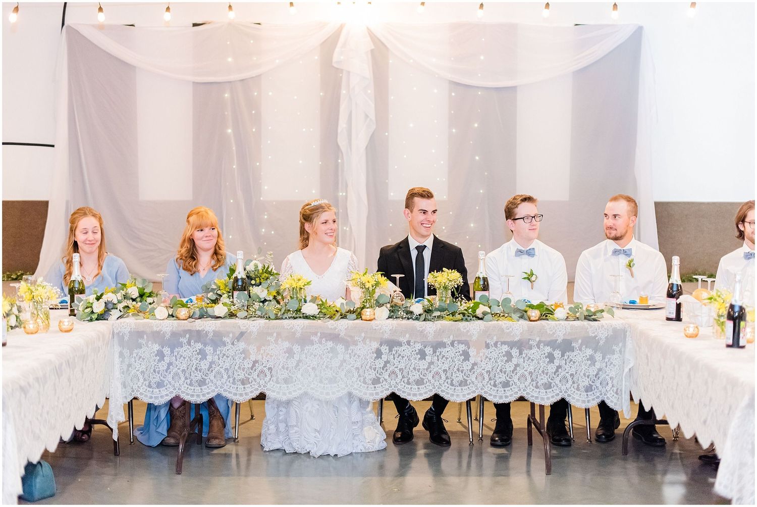 Wedding party sits at elegantly decorated head table with lace and greenery centerpieces.