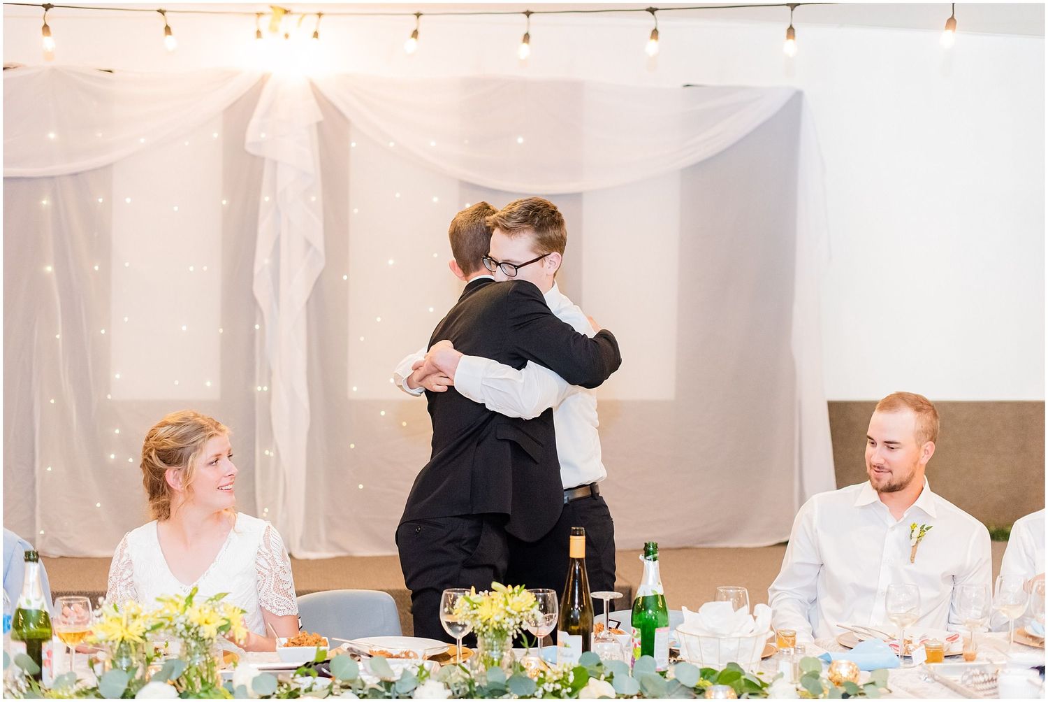Groom shares a heartfelt hug with a guest during wedding reception speeches while bride watches smiling nearby.