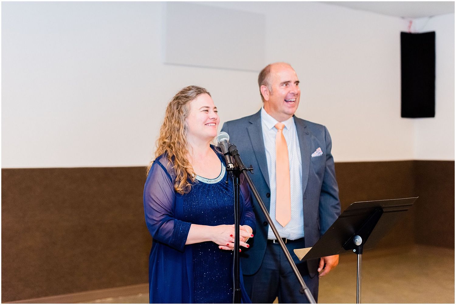 Couple in formal attire speaking at microphone during wedding reception toast in elegant venue.