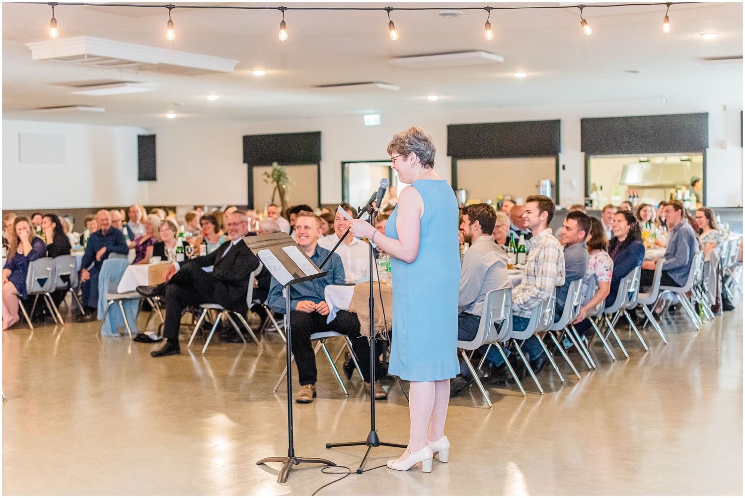 Female performer plays clarinet at wedding reception while guests watch from rows of chairs in bright modern venue.