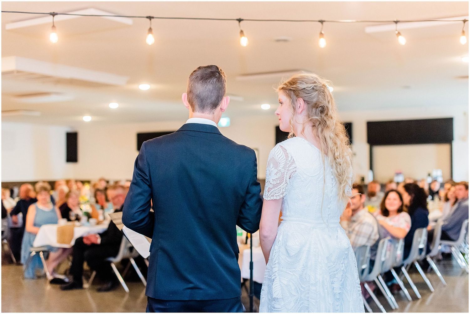 Bride and groom stand at a microphone addressing their wedding reception guests in a bright decorated venue.