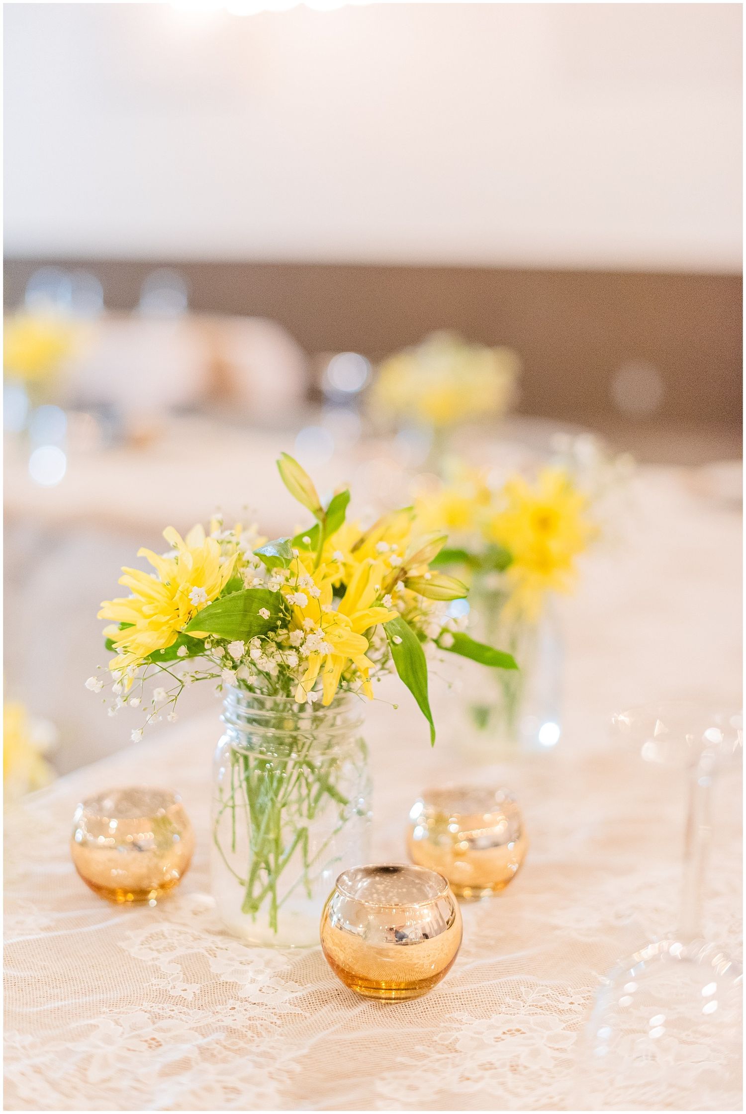 Yellow sunflowers and baby's breath in mason jar vases with gold mercury glass votives on lace table runners.