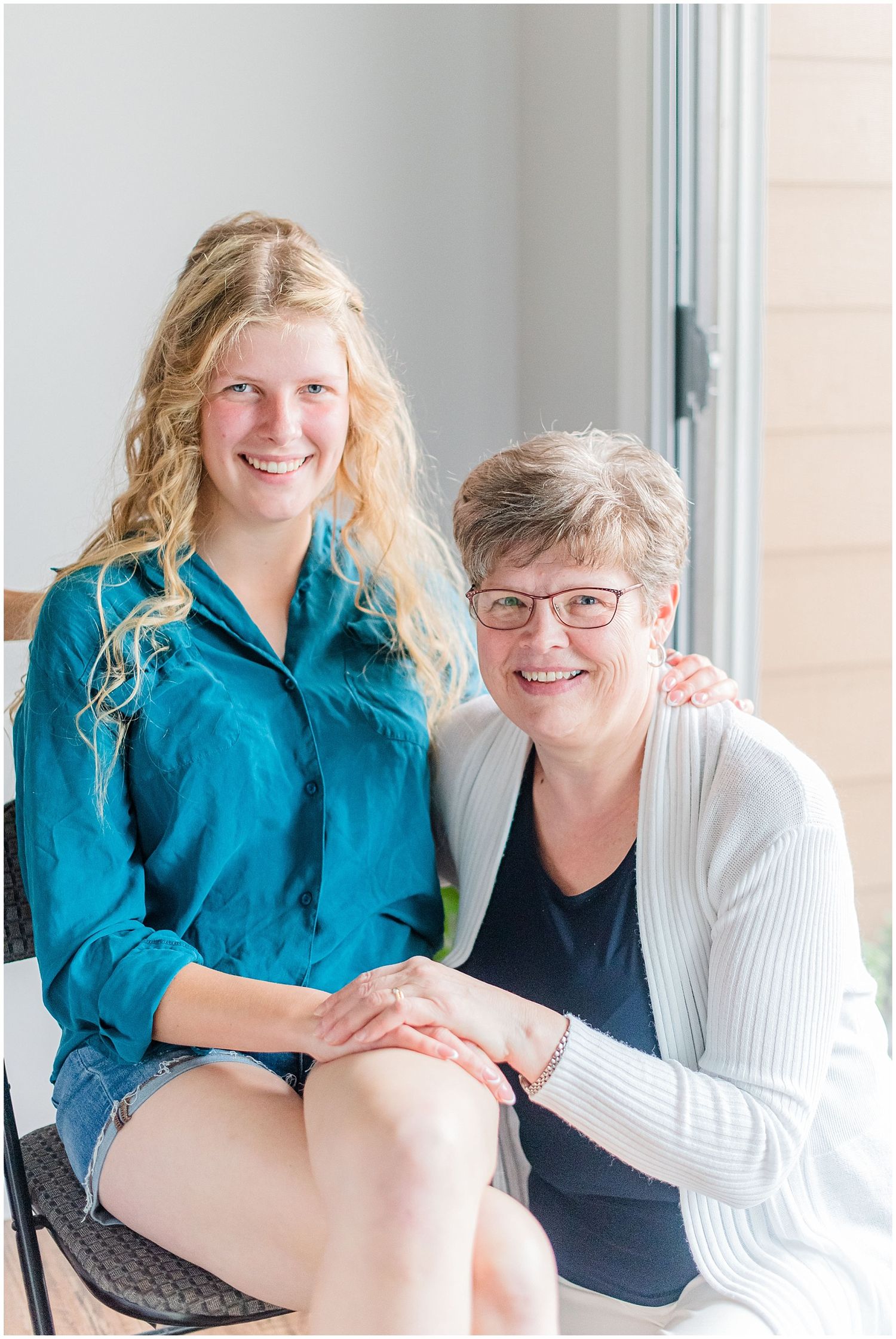 Two smiling women, one young with blonde hair in teal shirt and one older with glasses, pose together indoors.