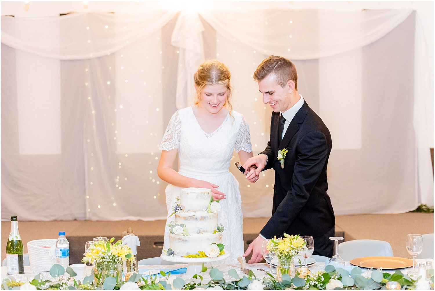 Bride and groom smile while cutting their white wedding cake at elegant decorated reception table.
