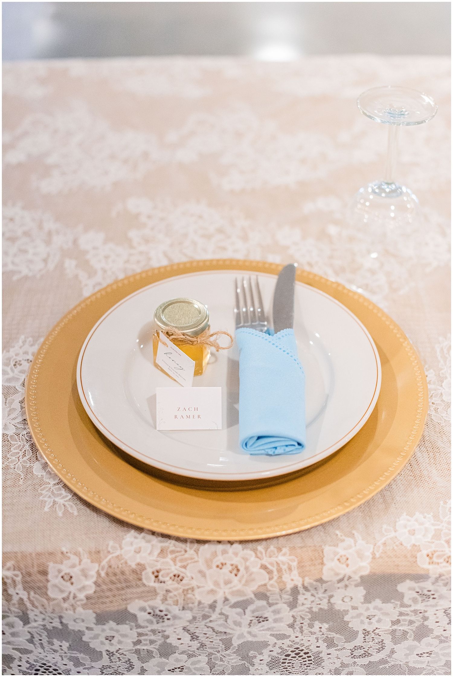 Gold charger plate with white china, blue napkin, honey jar favor, and place card on lace tablecloth.