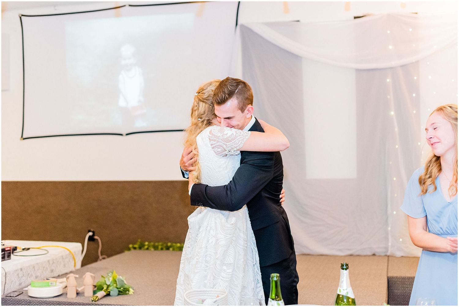 Bride and groom share a heartfelt hug during wedding reception while bridesmaid in blue dress looks on smiling.