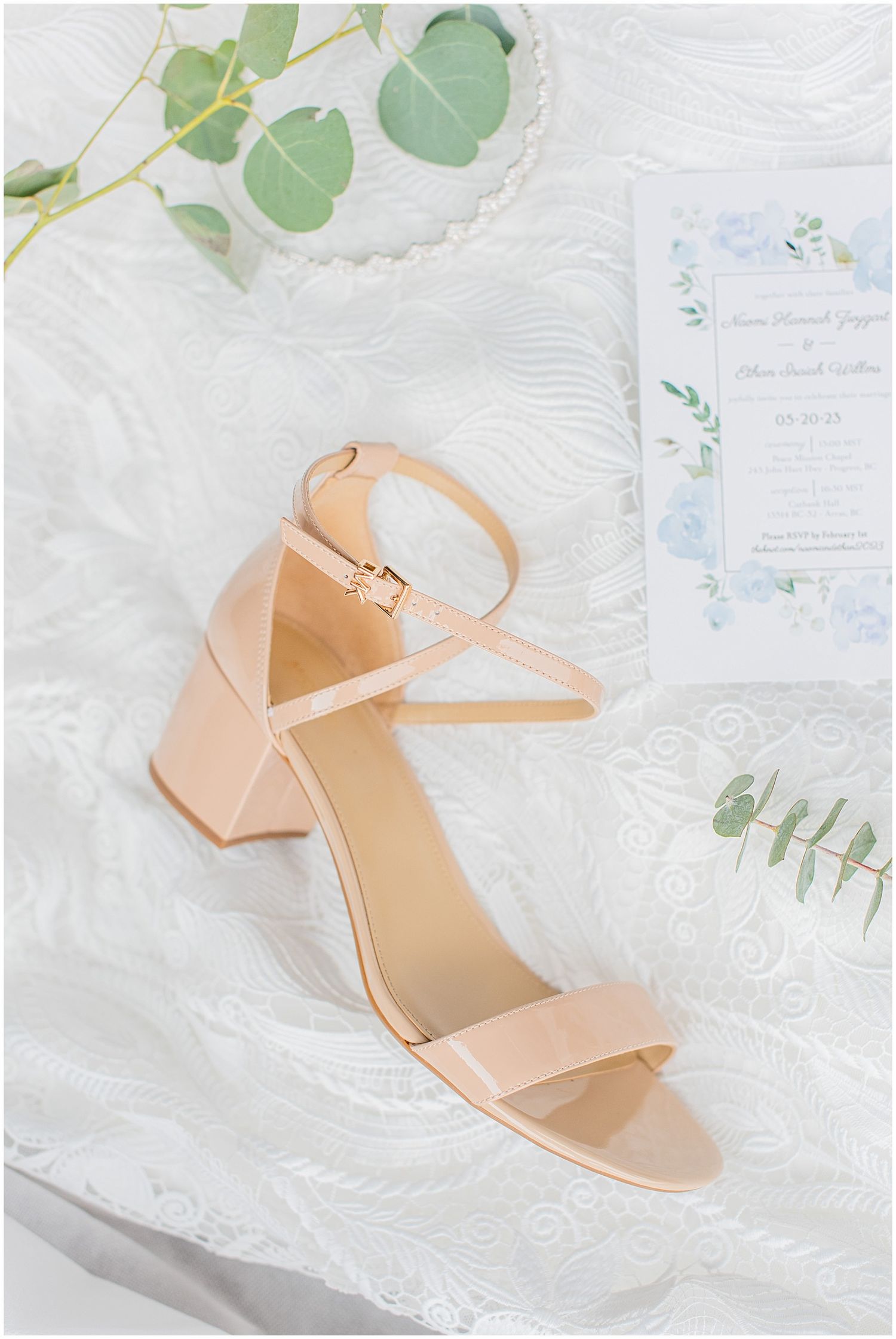 Blush pink block heel sandal displayed on white lace fabric beside a floral wedding invitation and eucalyptus greenery.