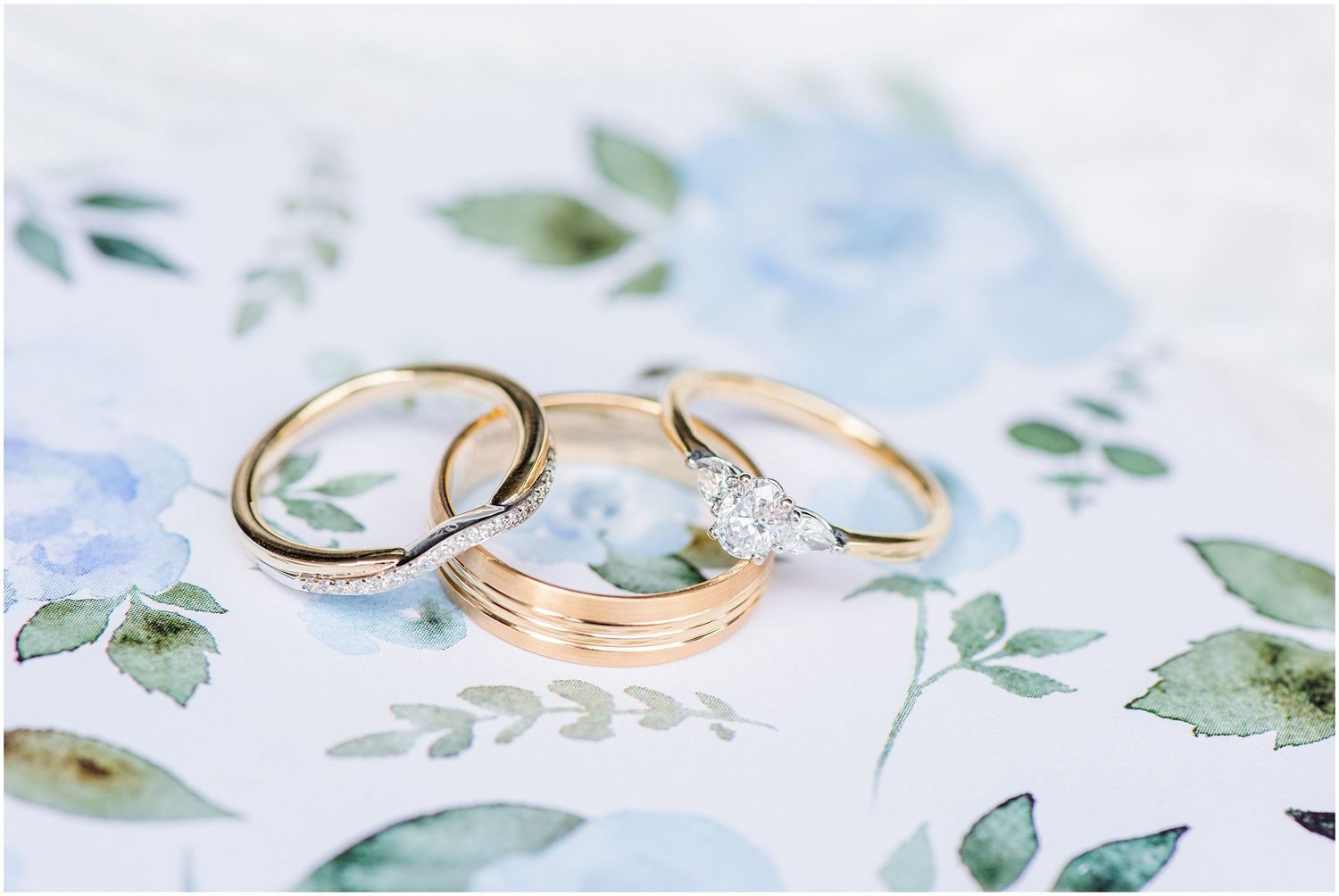 Three gold wedding and engagement rings with diamond solitaire displayed on floral watercolor background.