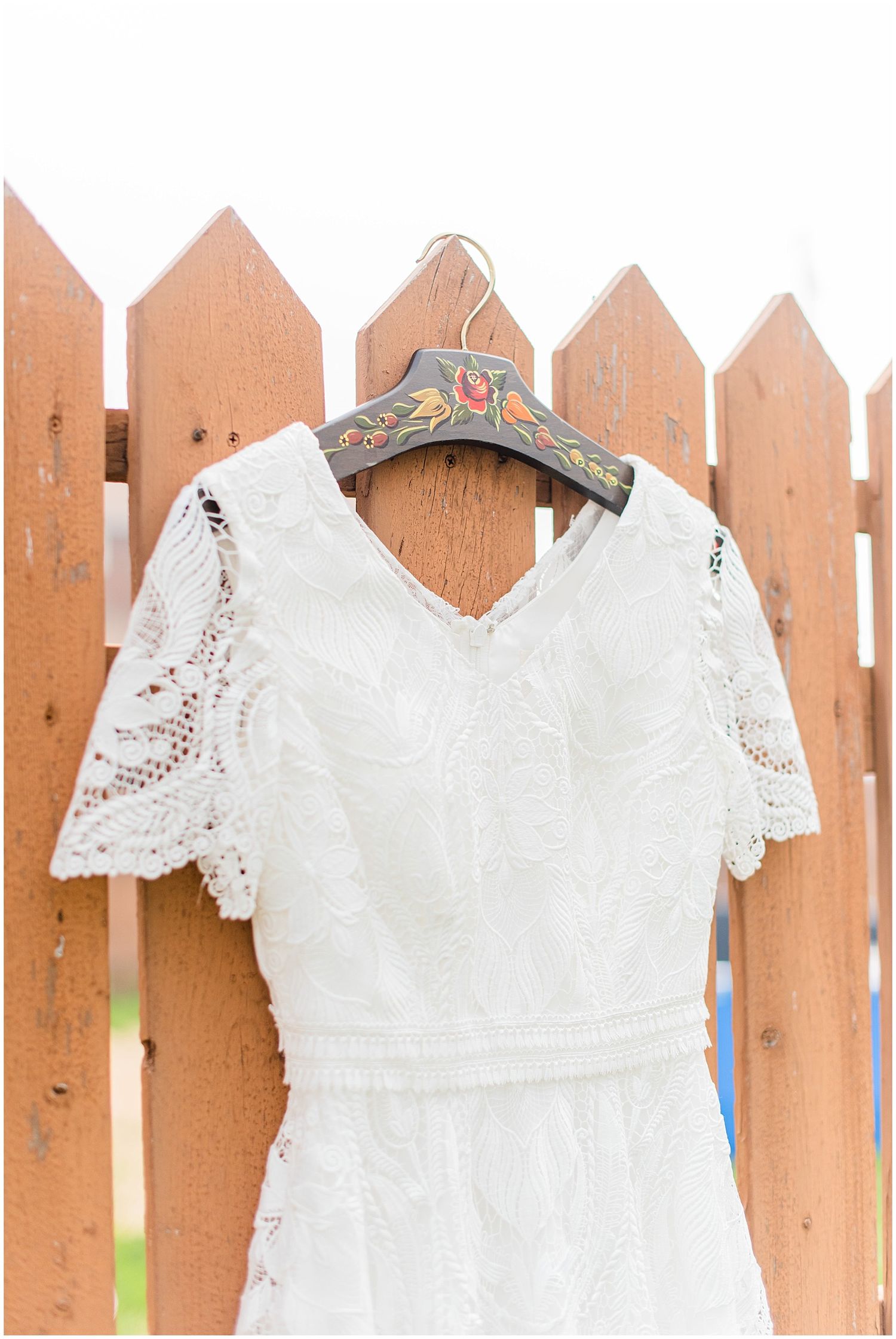 White lace wedding dress with flutter sleeves hanging on decorated hanger on rustic wooden fence.