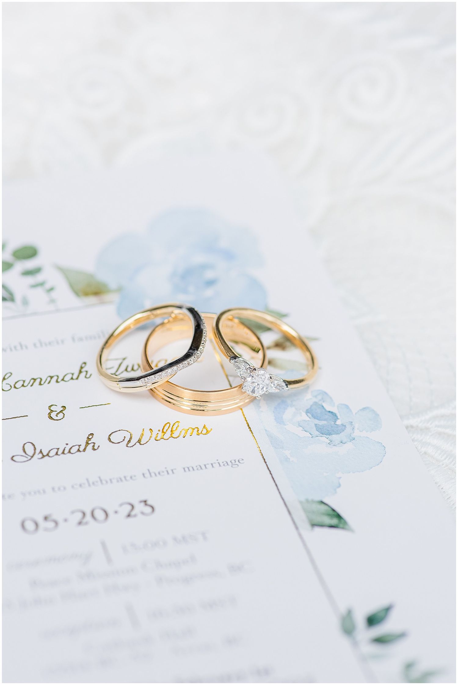 Gold wedding bands and diamond engagement ring resting on floral wedding invitation dated 05-20-23.