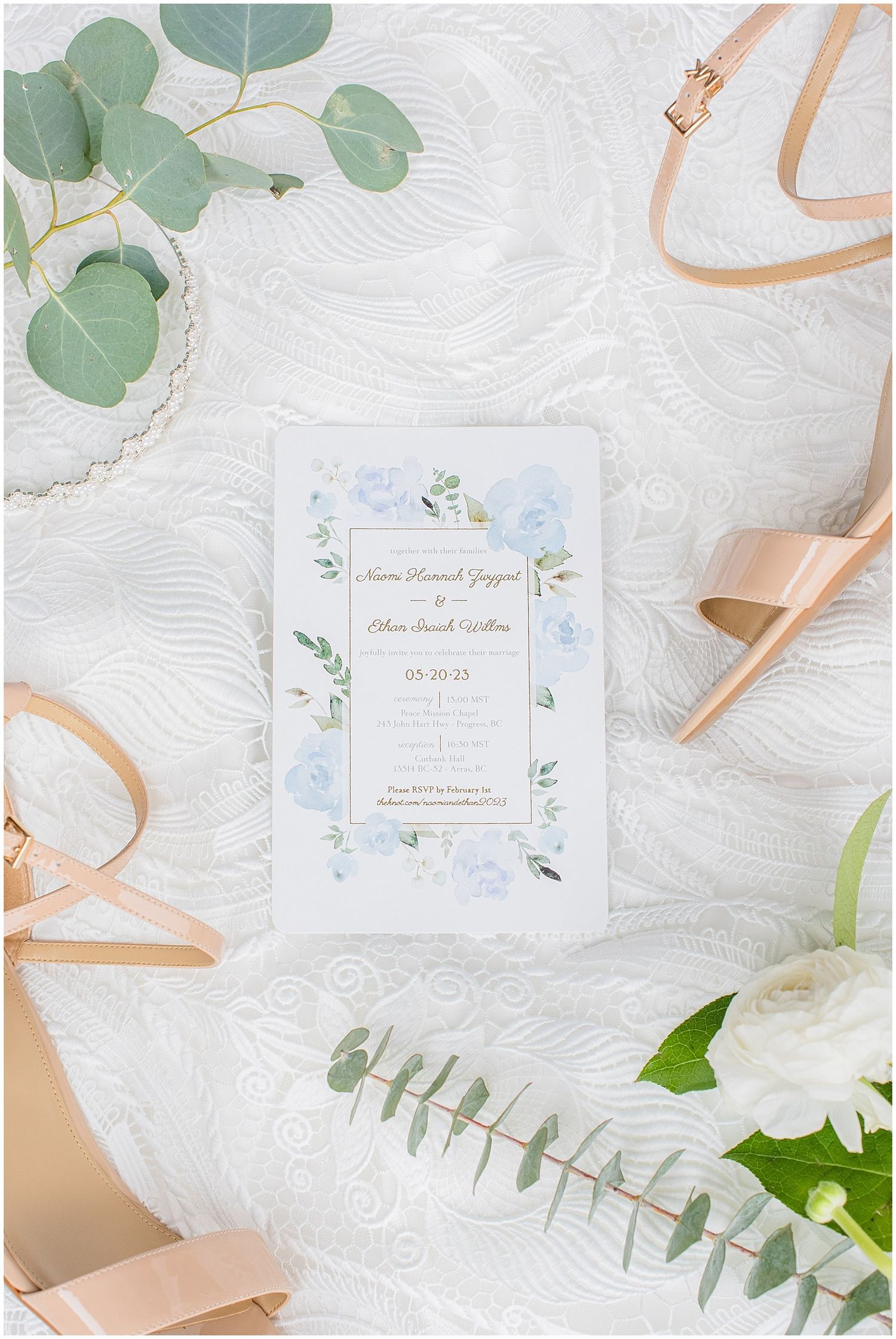 Elegant wedding invitation with blue floral design surrounded by nude heels, eucalyptus, and white flowers on lace fabric.