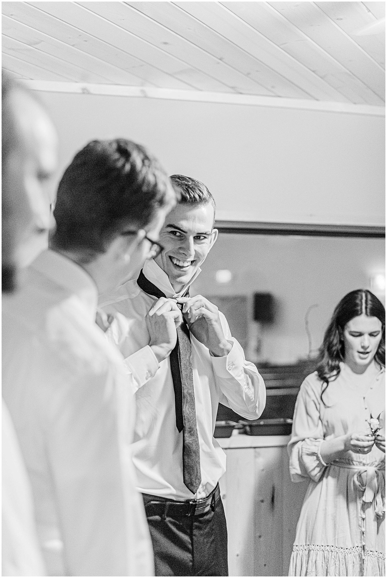 Smiling groom adjusting his tie while groomsmen prepare nearby in a bright indoor venue.