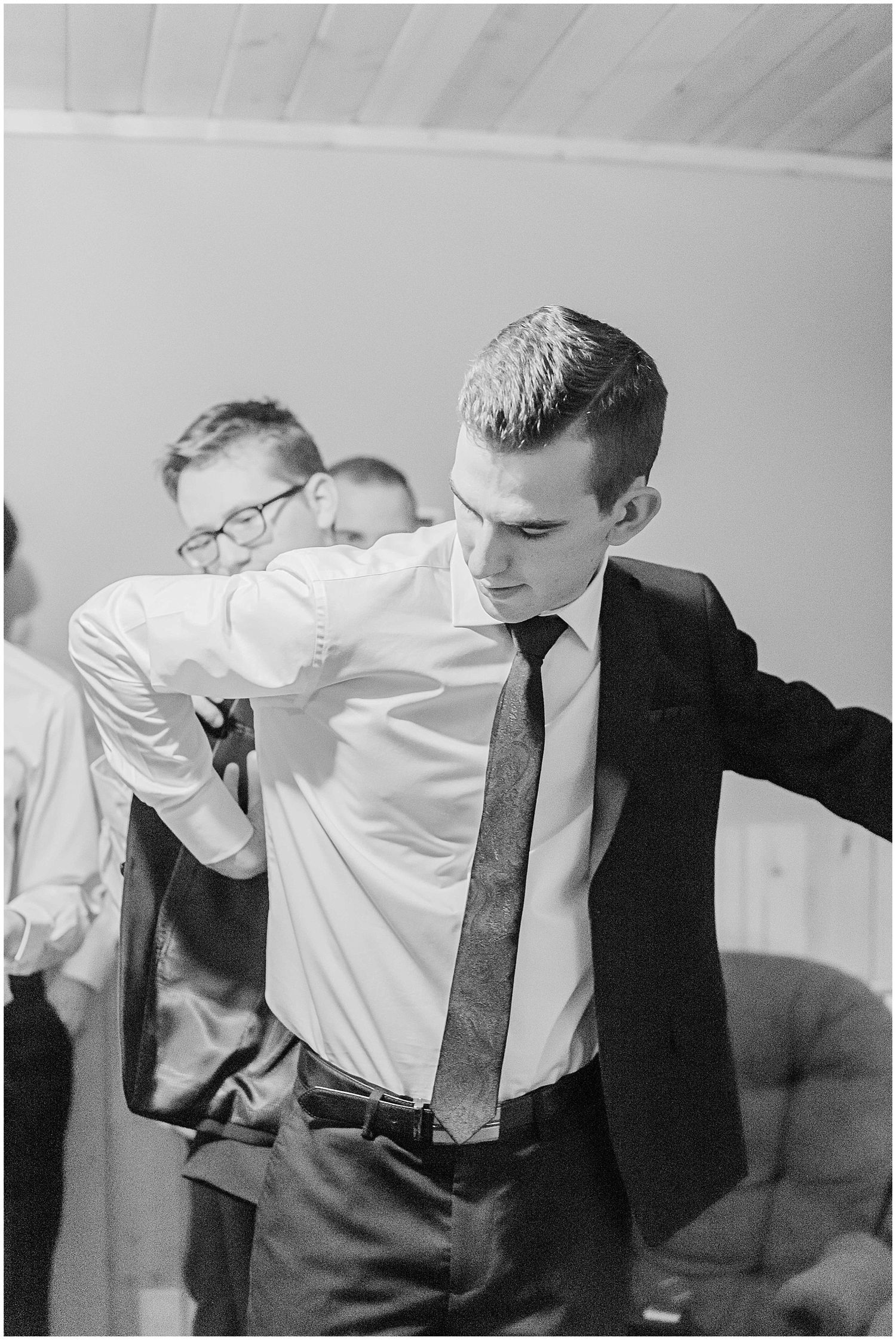 Groomsman being helped into his suit jacket by friends in black and white photo during wedding day preparations.