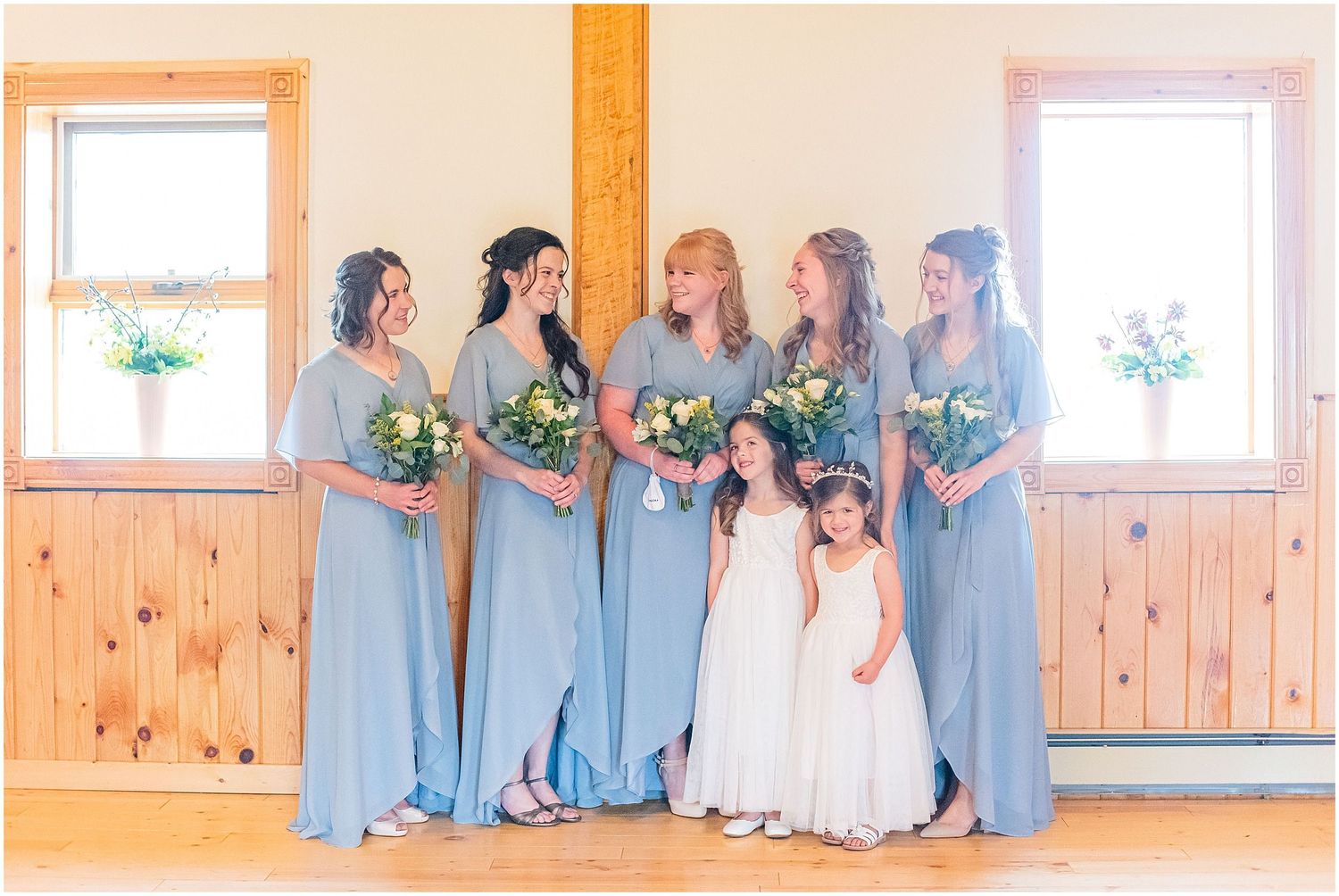 Bridesmaids in blue dresses holding floral bouquets with flower girls in white in a rustic venue.
