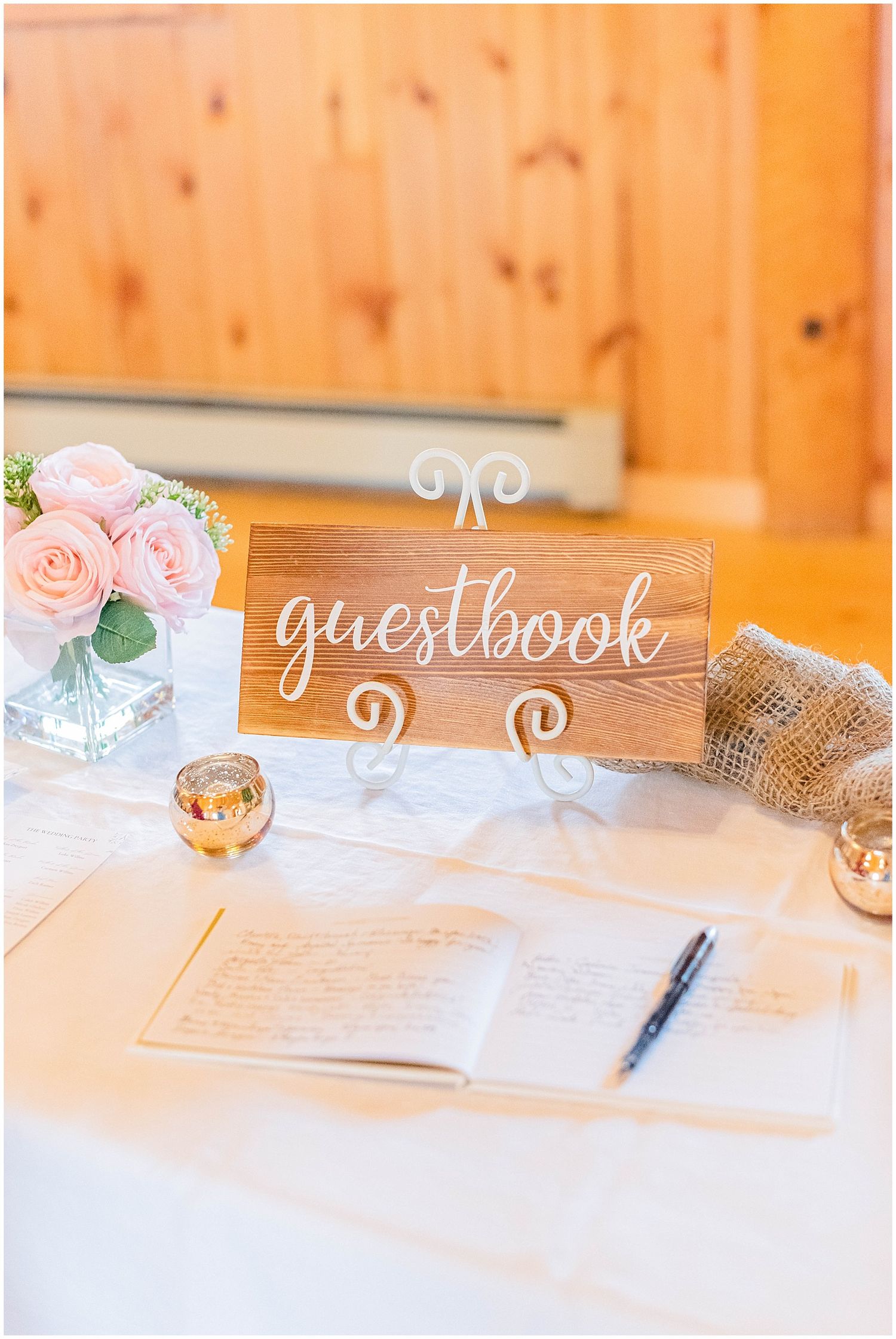 Rustic wooden guestbook sign on white table with pink roses and open book at wedding reception.
