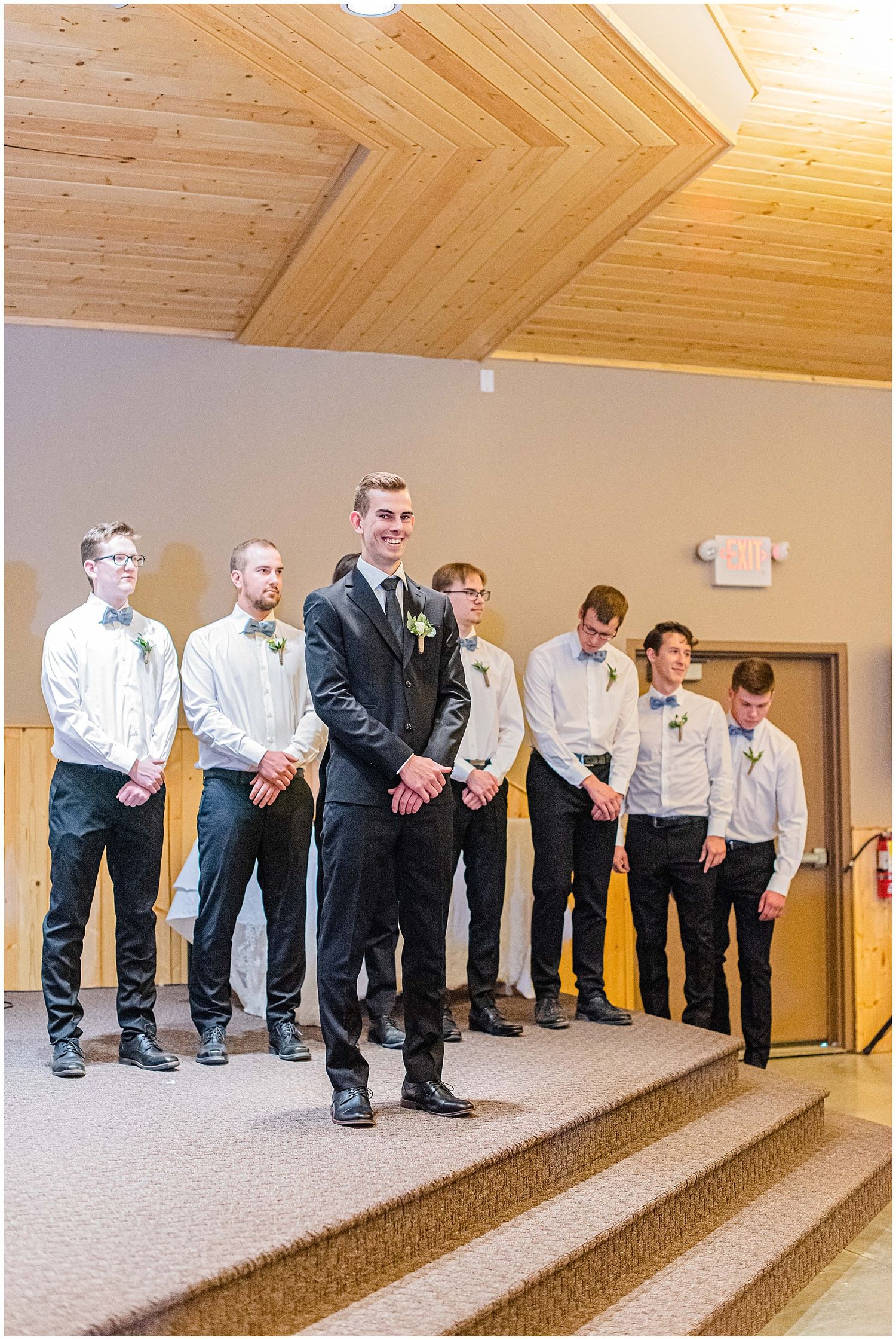 Groom in black suit stands at church altar with groomsmen in white shirts and black pants during wedding ceremony.