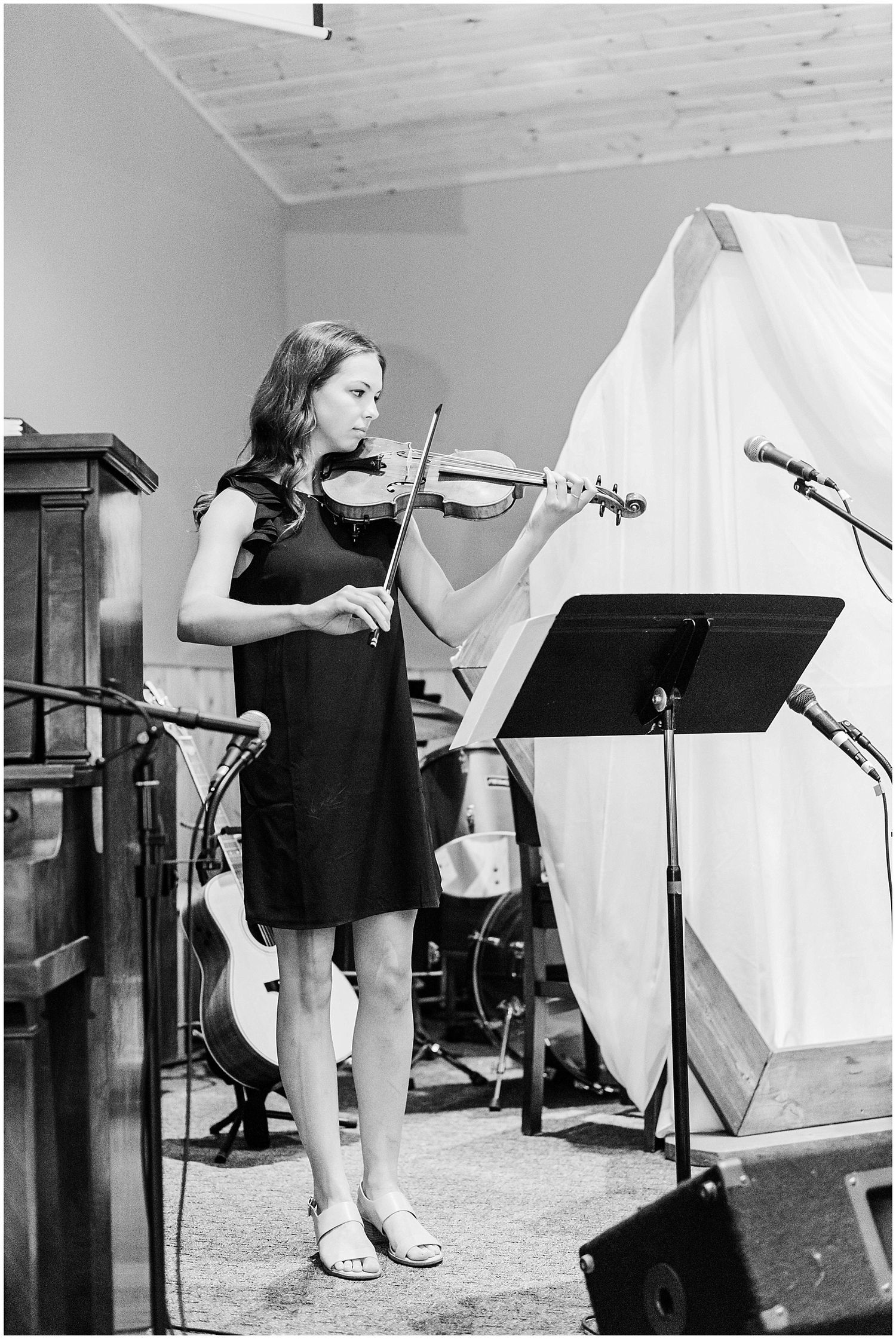 Black and white photo of young woman playing violin at wedding ceremony with music stand and instruments nearby.