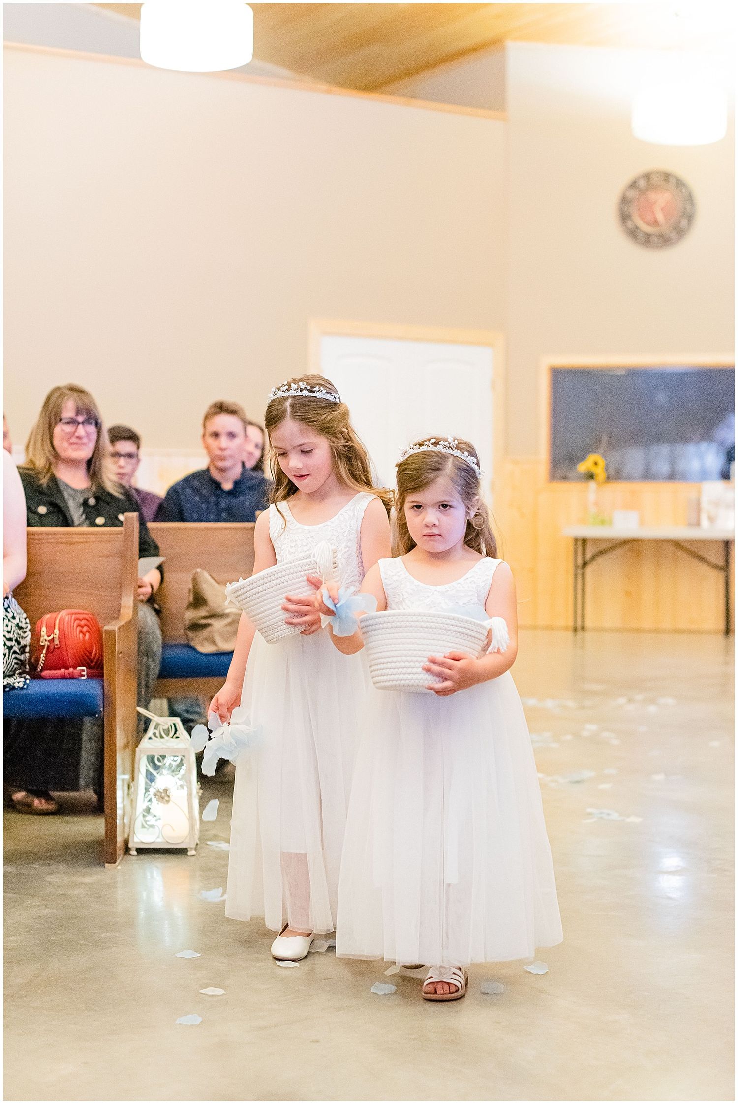 Two flower girls in white dresses and tiaras walk down church aisle with baskets.