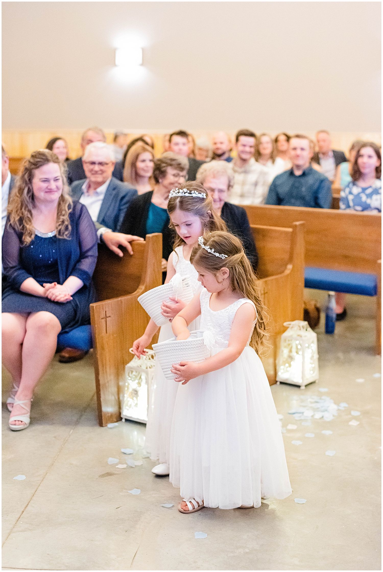 Two adorable flower girls in white dresses scatter petals down church aisle at wedding.