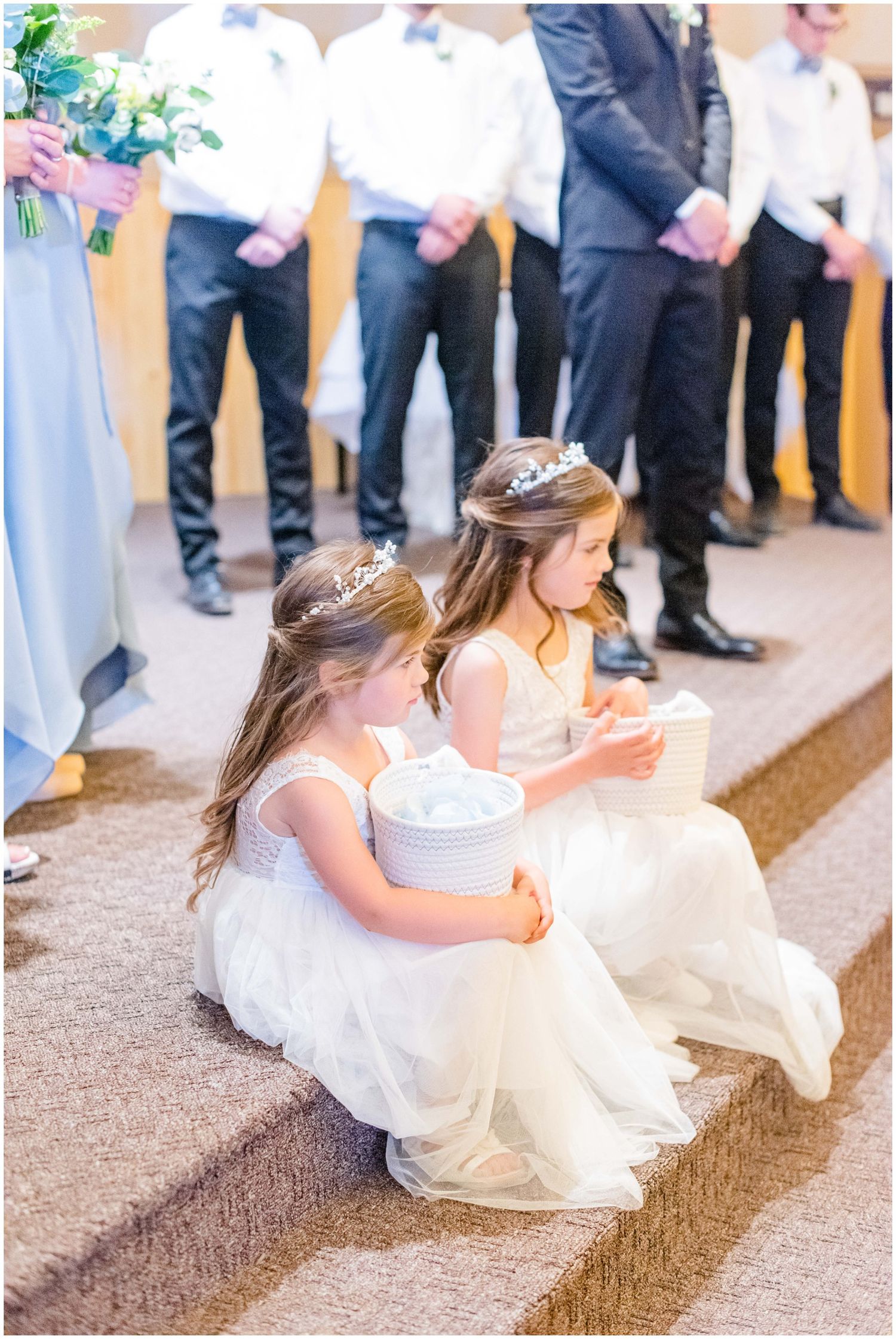 Two flower girls in white dresses sitting on church altar steps holding baskets during wedding ceremony.