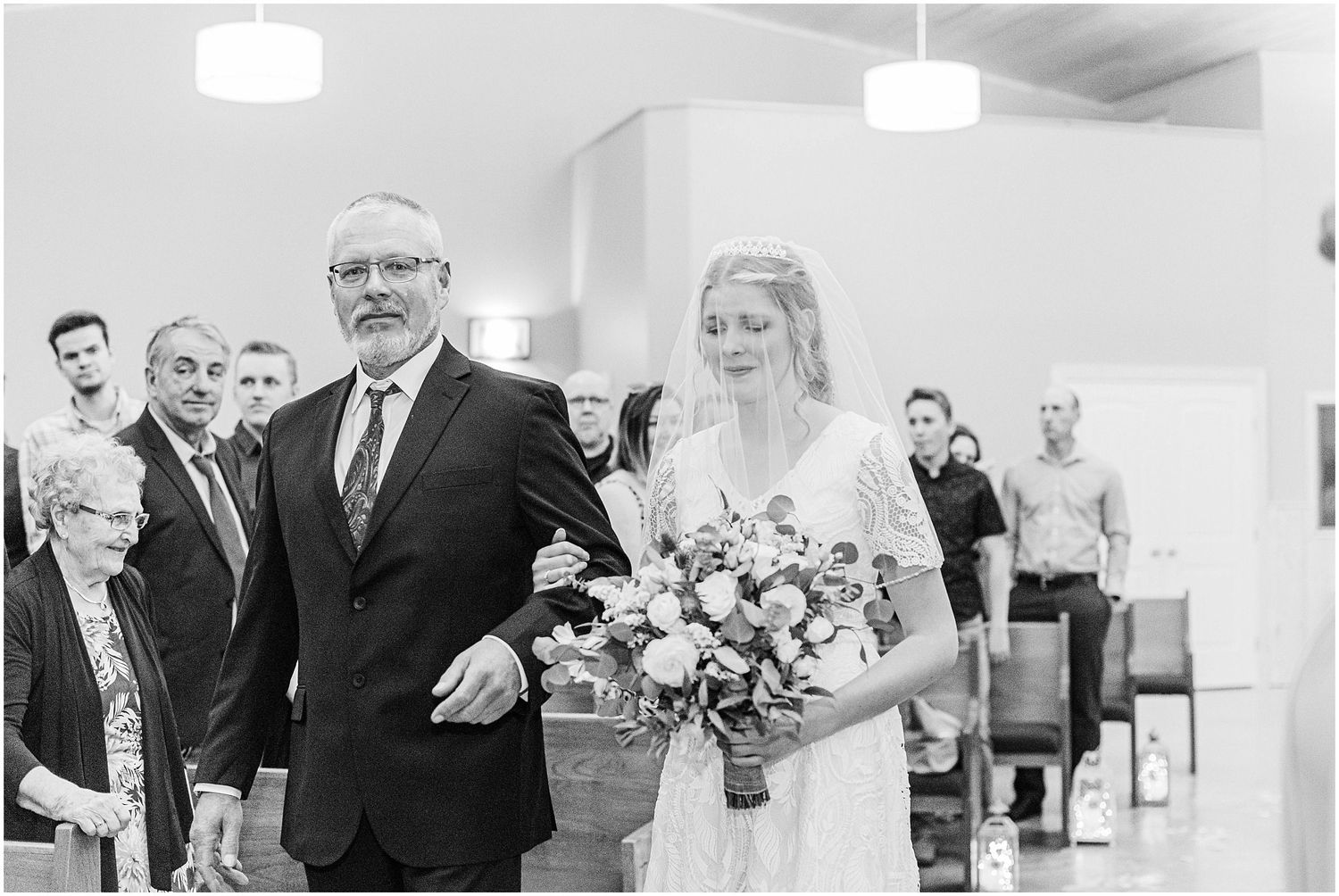 Black and white photo of bride walking down church aisle escorted by her father holding a bouquet.