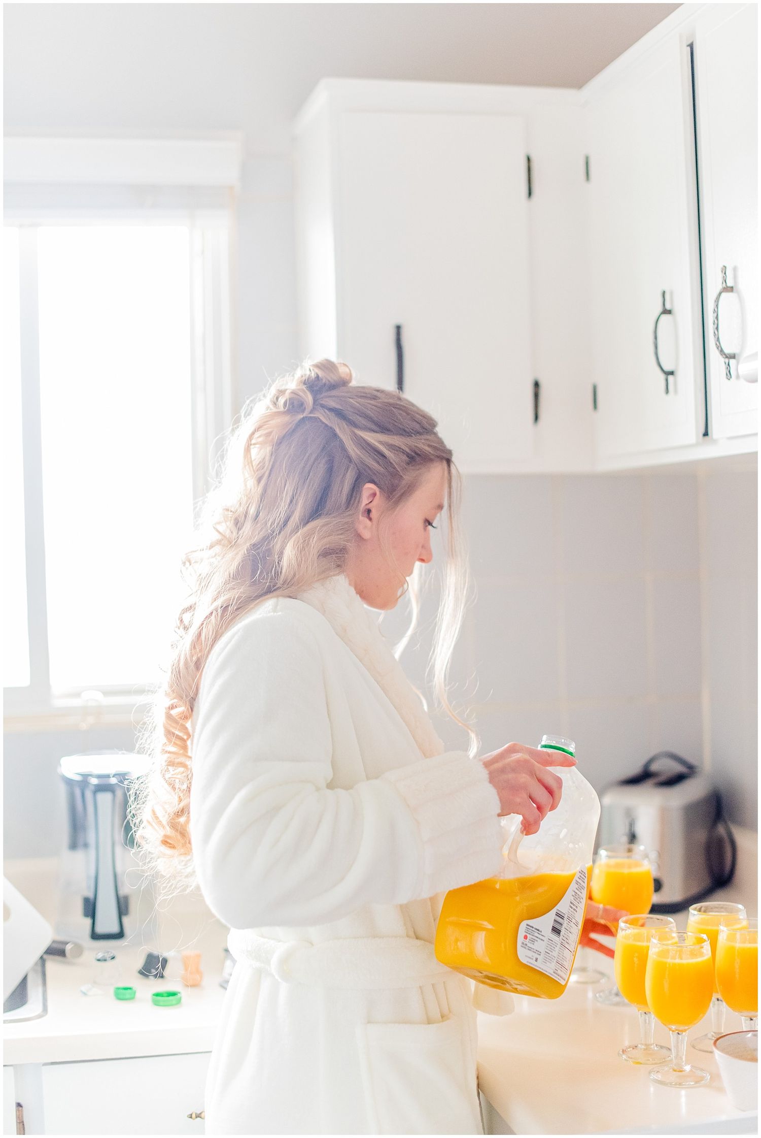 Bride in white robe pouring orange juice for mimosas in bright white kitchen during wedding morning preparations.