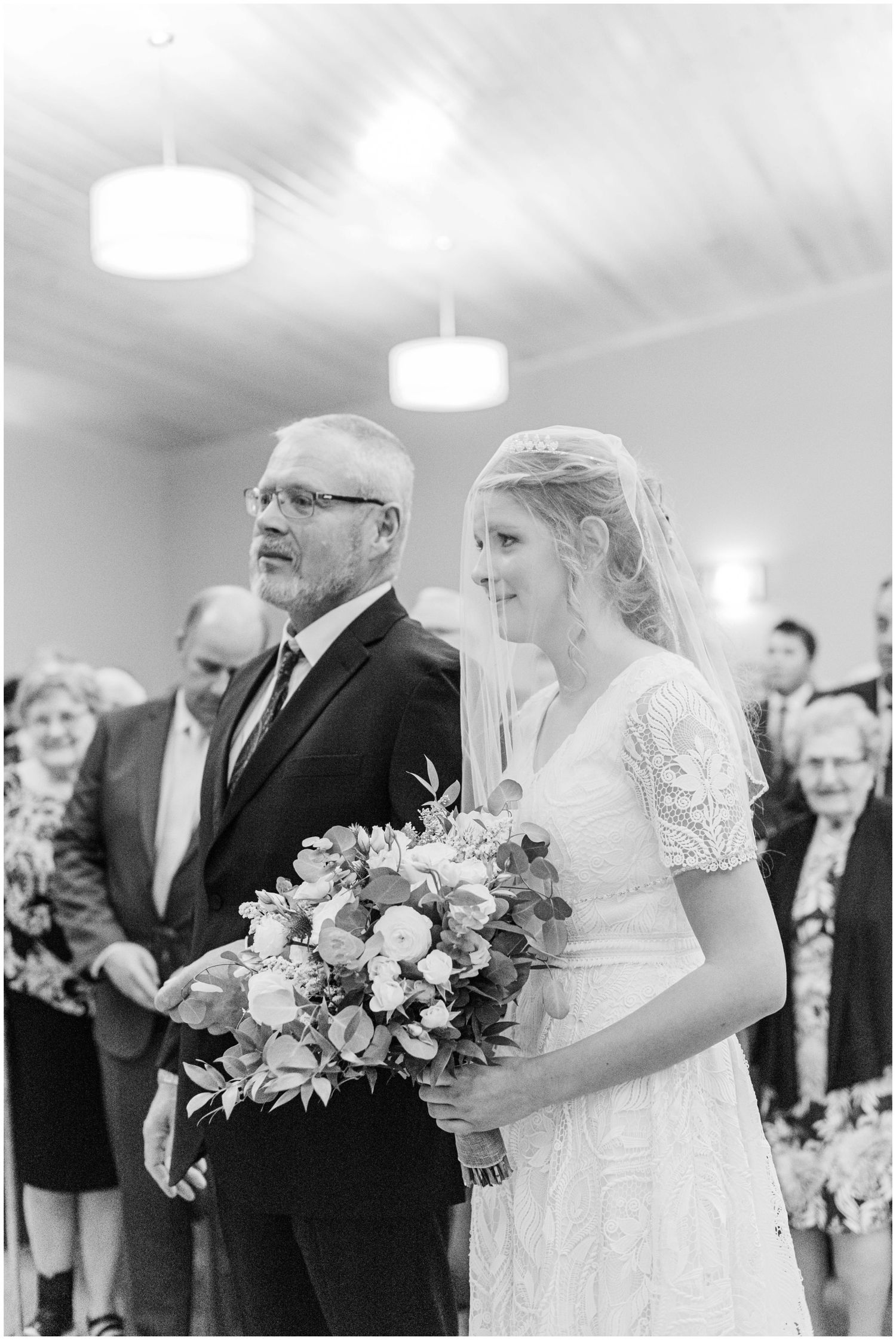 Bride holding a large floral bouquet walks down the aisle with her father during a wedding ceremony.