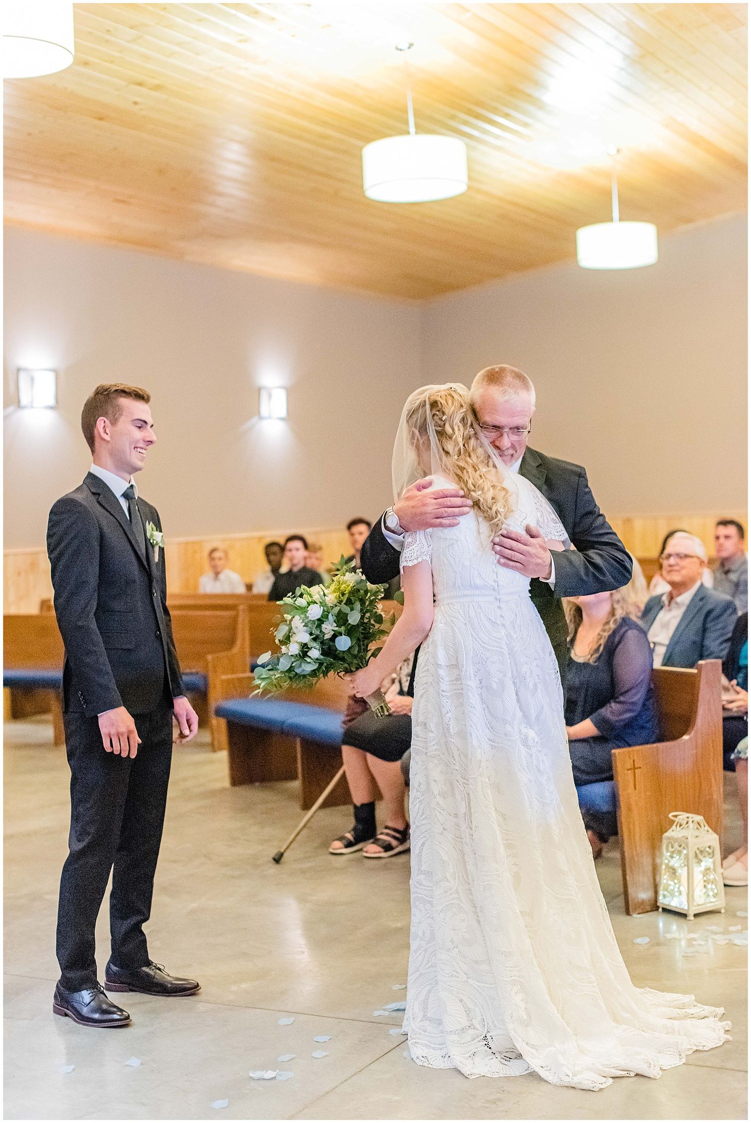 Bride hugging her father at the altar during a wedding ceremony in a chapel, with the groom standing nearby.