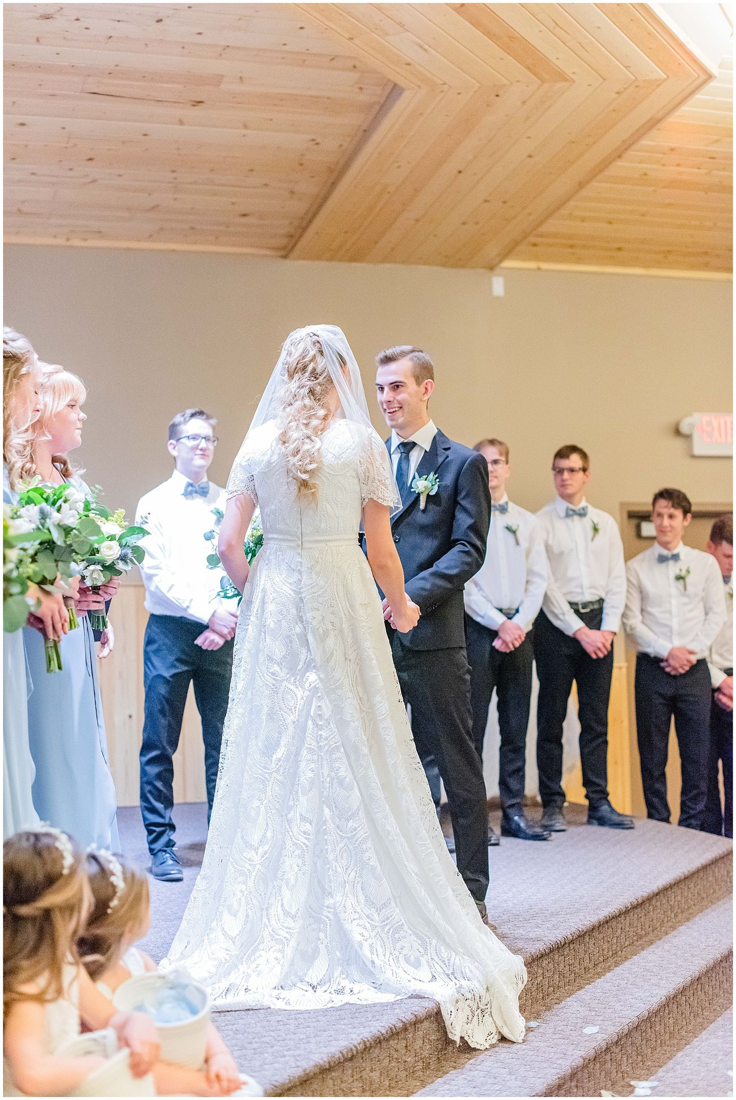 Bride and groom stand at the altar during wedding ceremony, surrounded by bridesmaids and groomsmen in a wooden chapel.
