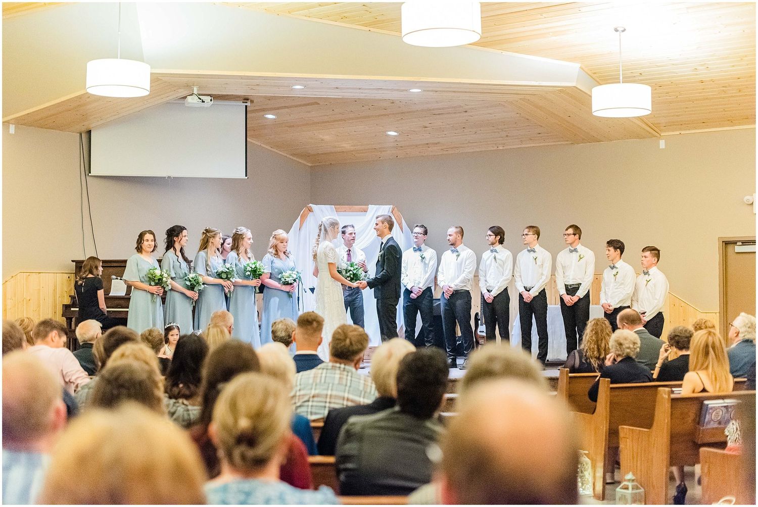 Wedding party stands at church altar with bridesmaids in grey and groomsmen in white during ceremony.