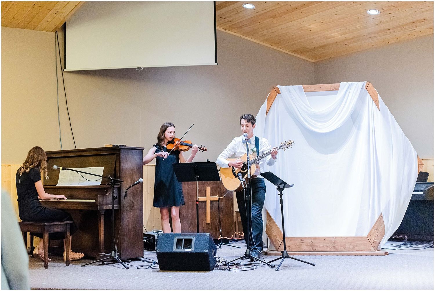 Musicians perform at a church wedding ceremony with violin, guitar, and piano on a wooden stage.