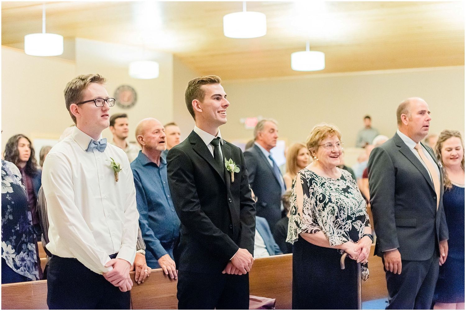 Groom stands at the altar with groomsmen and family smiling during a wedding ceremony in a church.