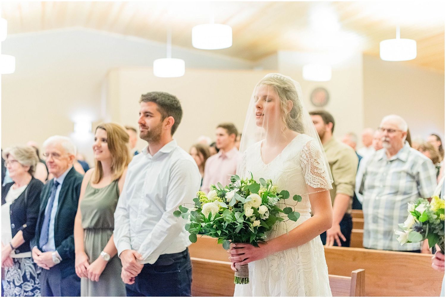 Bride holding white and green bouquet stands with guests in church pews during a wedding ceremony.