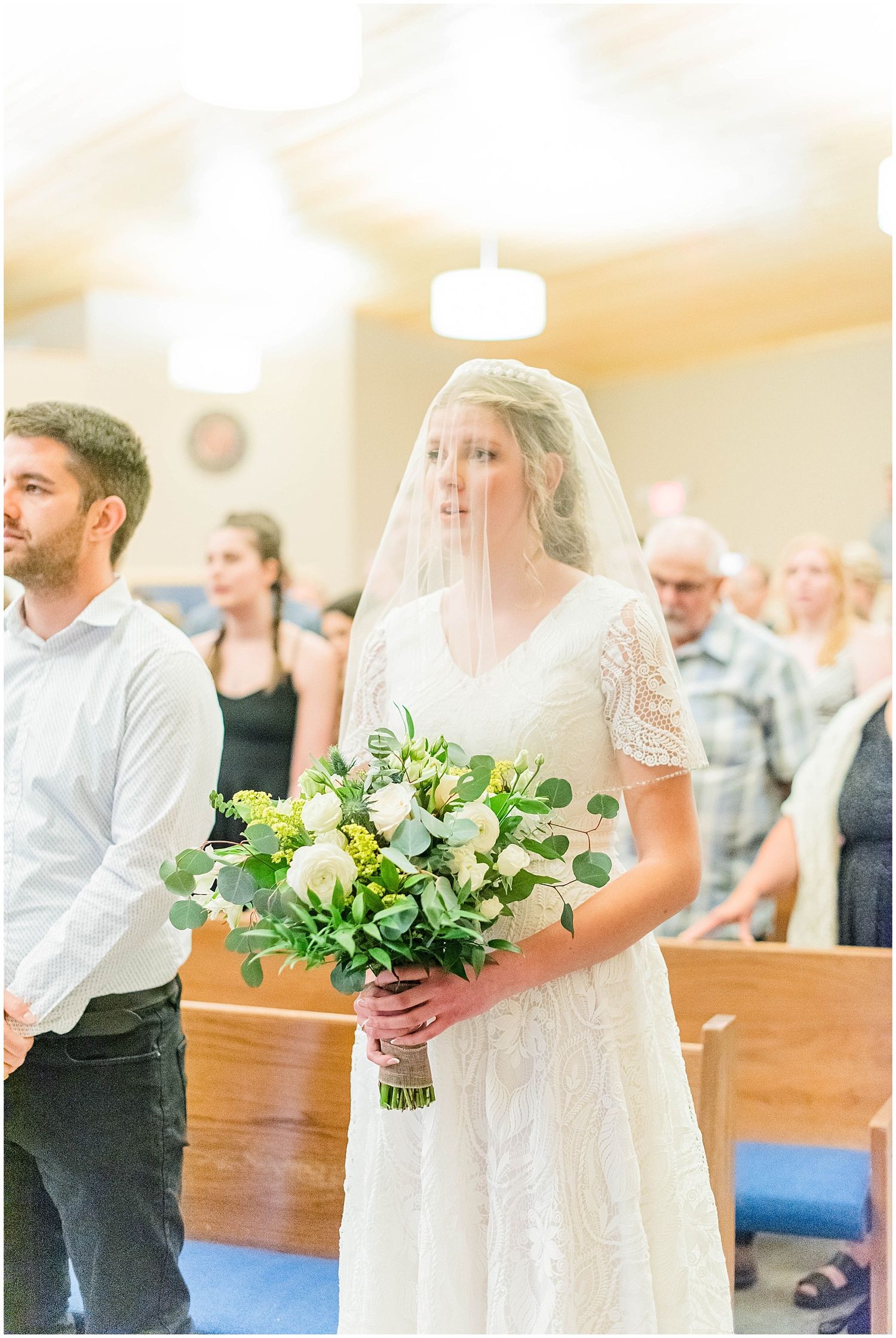 Bride walks down church aisle in lace gown with veil holding white and green bouquet.