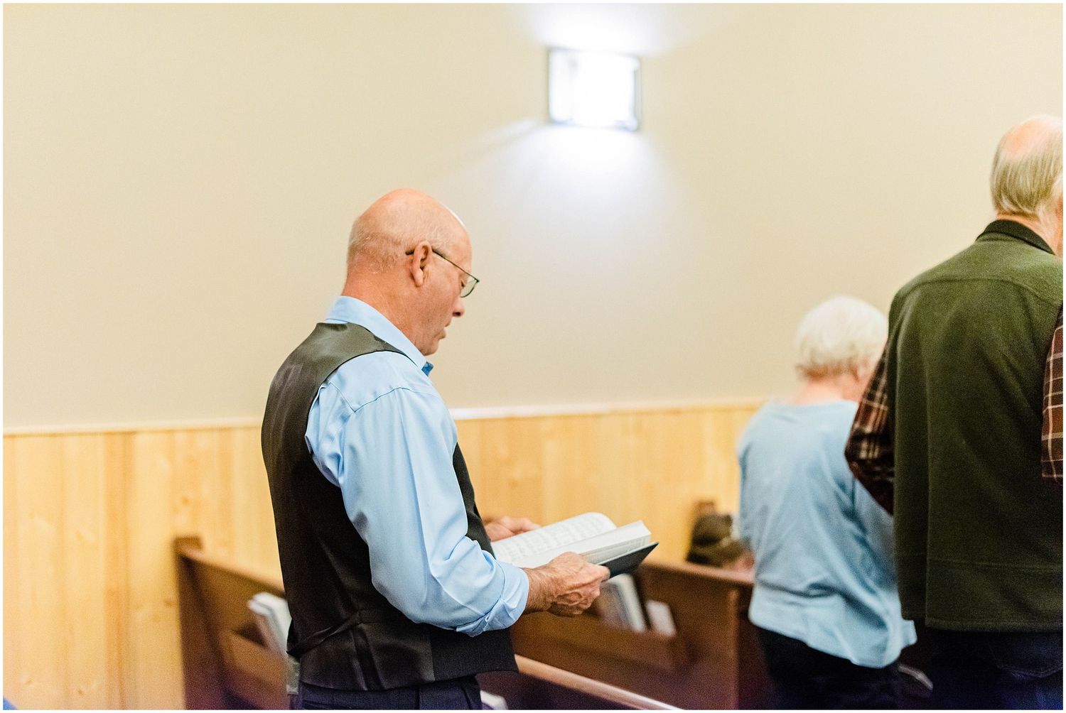 Bald man with glasses reading from a book while standing in a wooden-paneled church pew.