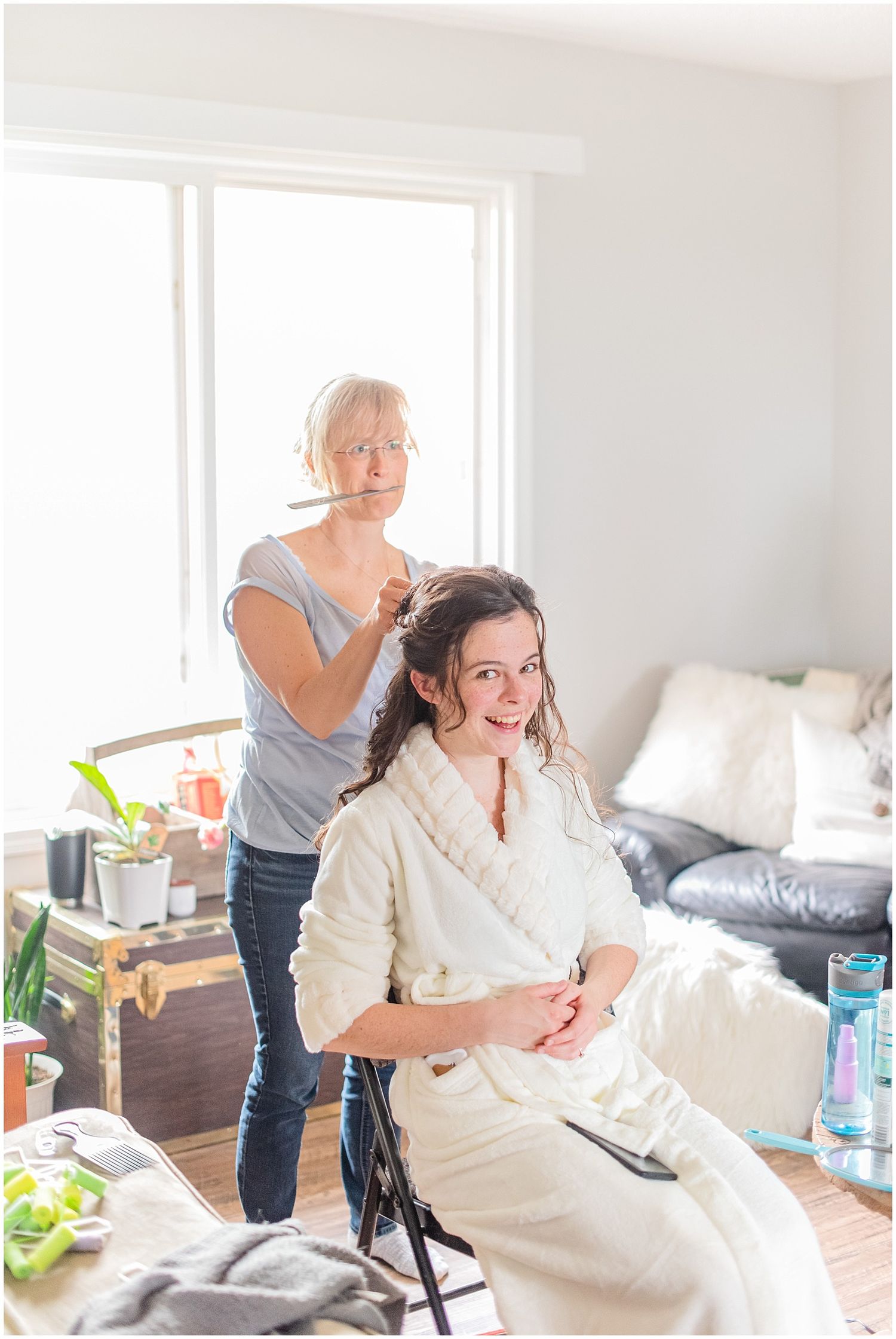 A hairstylist works on a smiling bride wearing a white robe while she sits in a bright, cozy room before her wedding.