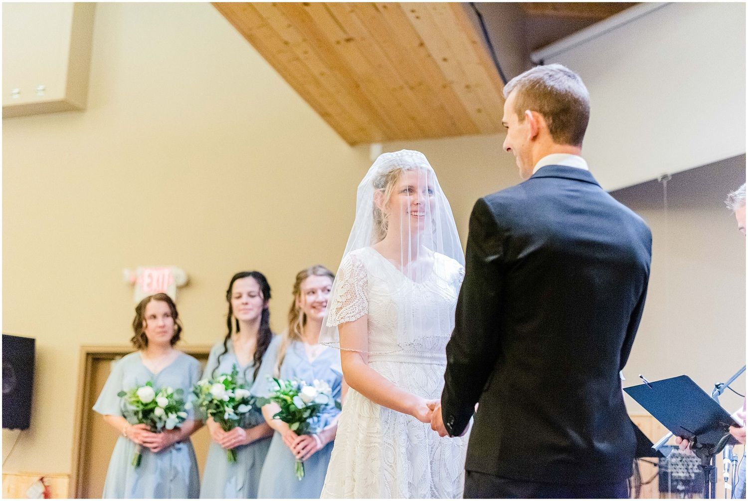 A bride in a lace gown and veil holds hands with the groom at the altar flanked by bridesmaids.