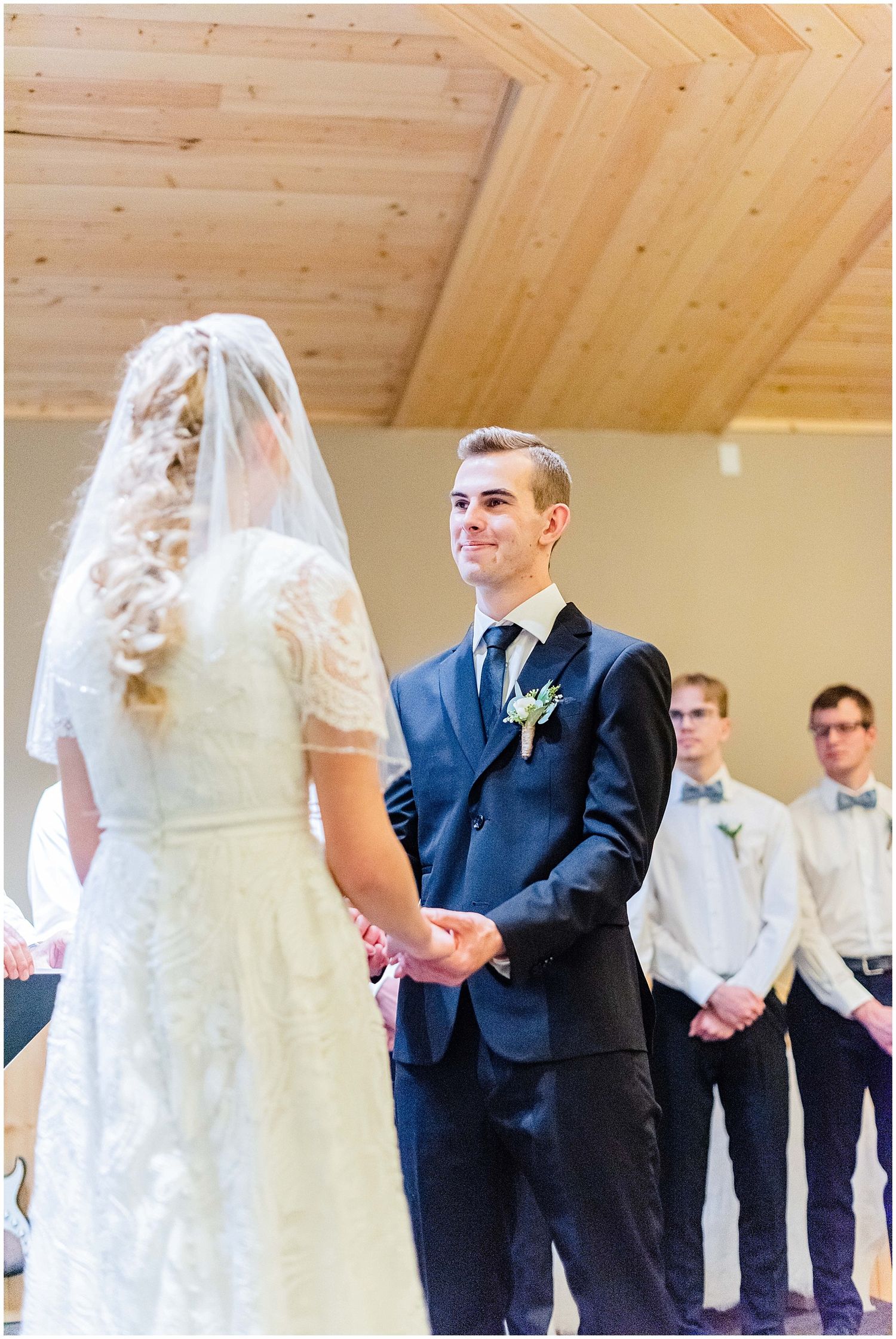 Groom smiling at bride while holding hands during indoor wedding ceremony with groomsmen standing in background.