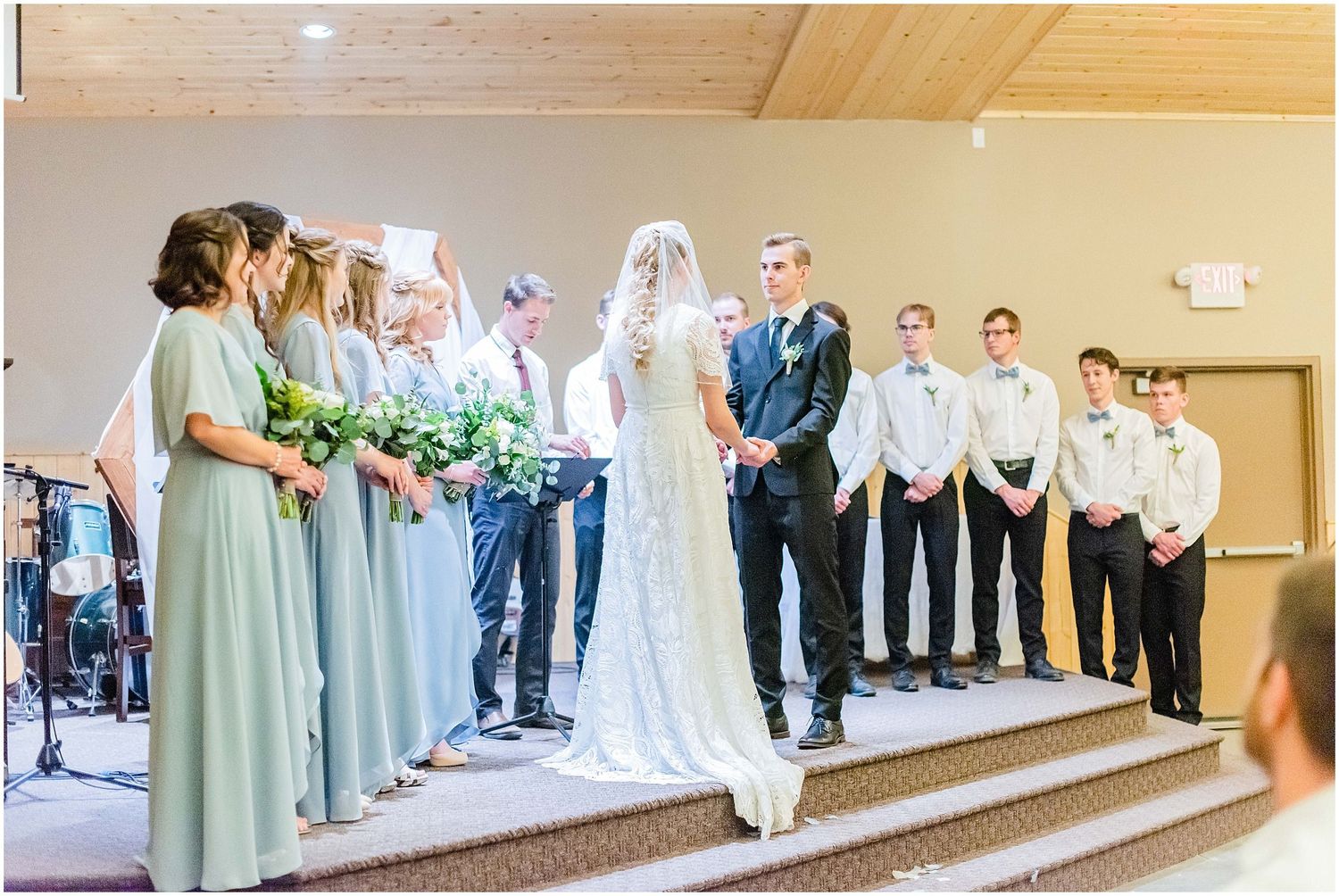 Bride and groom stand at the altar surrounded by bridesmaids in sage green dresses and groomsmen.