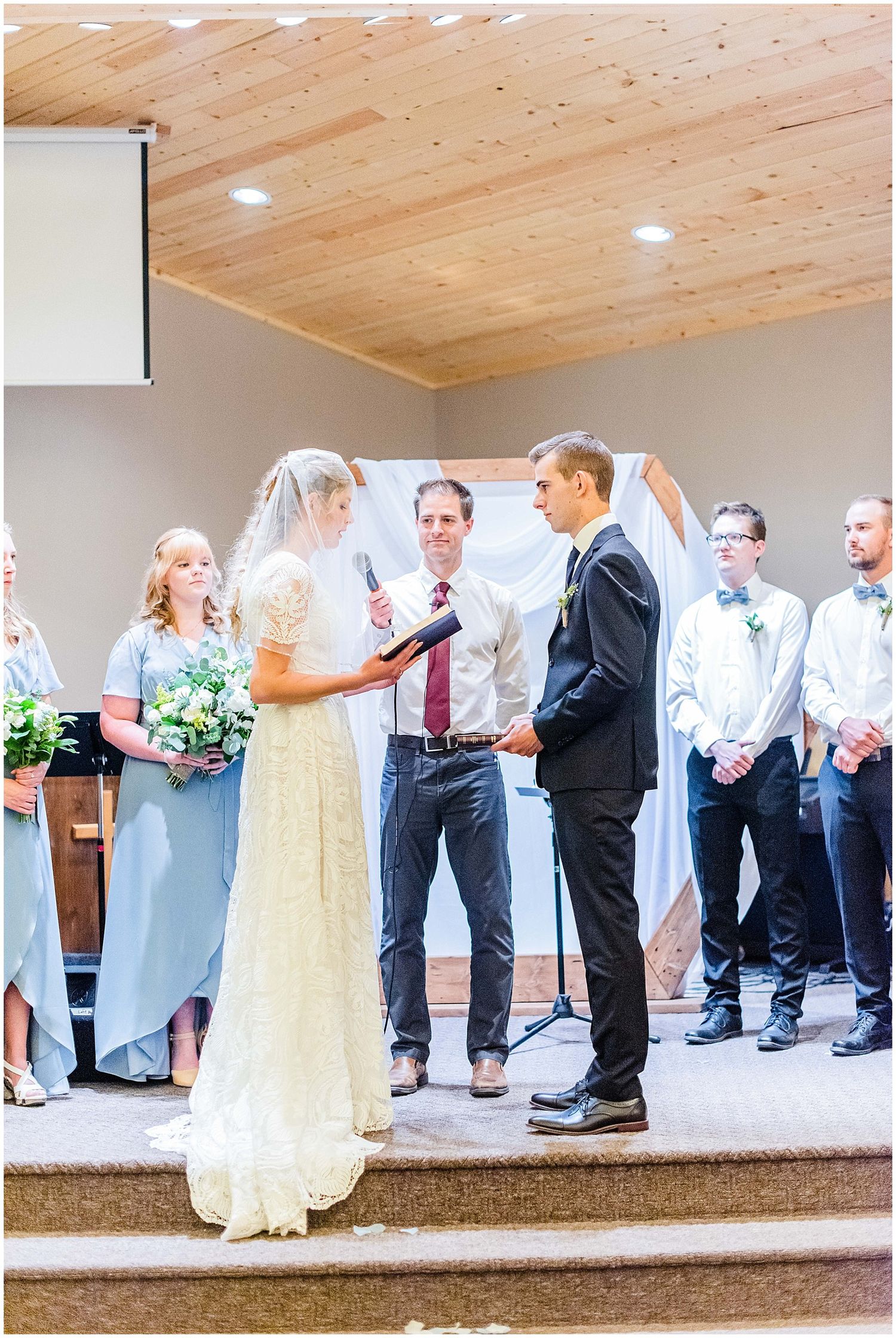 Bride and groom exchange vows at church altar with bridesmaids in blue dresses and groomsmen in black suits.