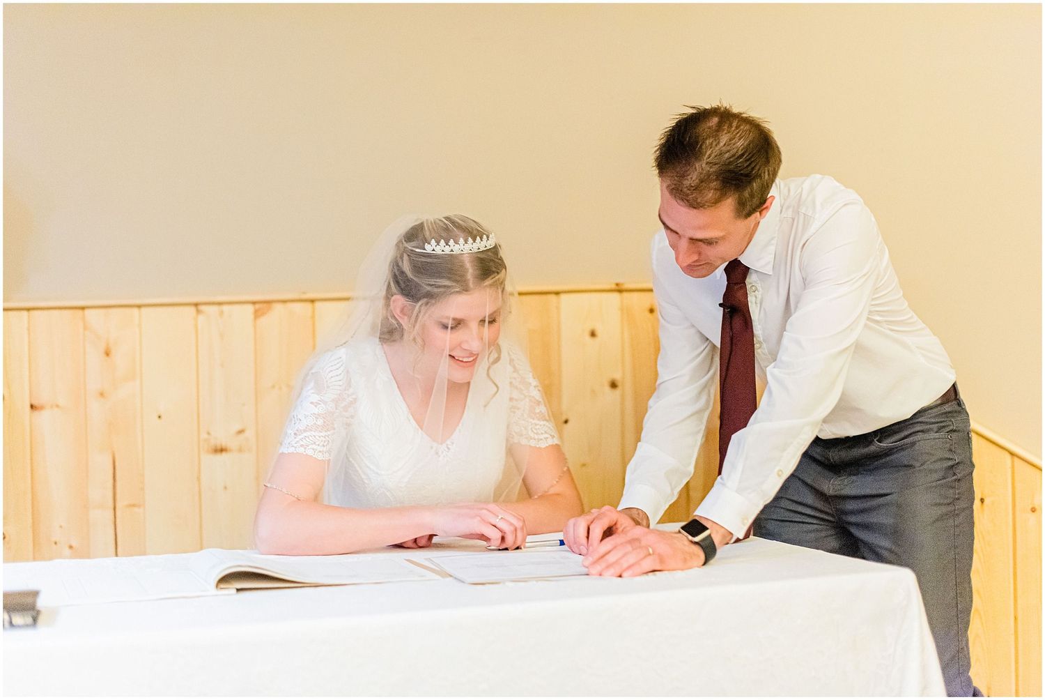 Bride and groom signing wedding registry documents at a white table in a wood-paneled room.