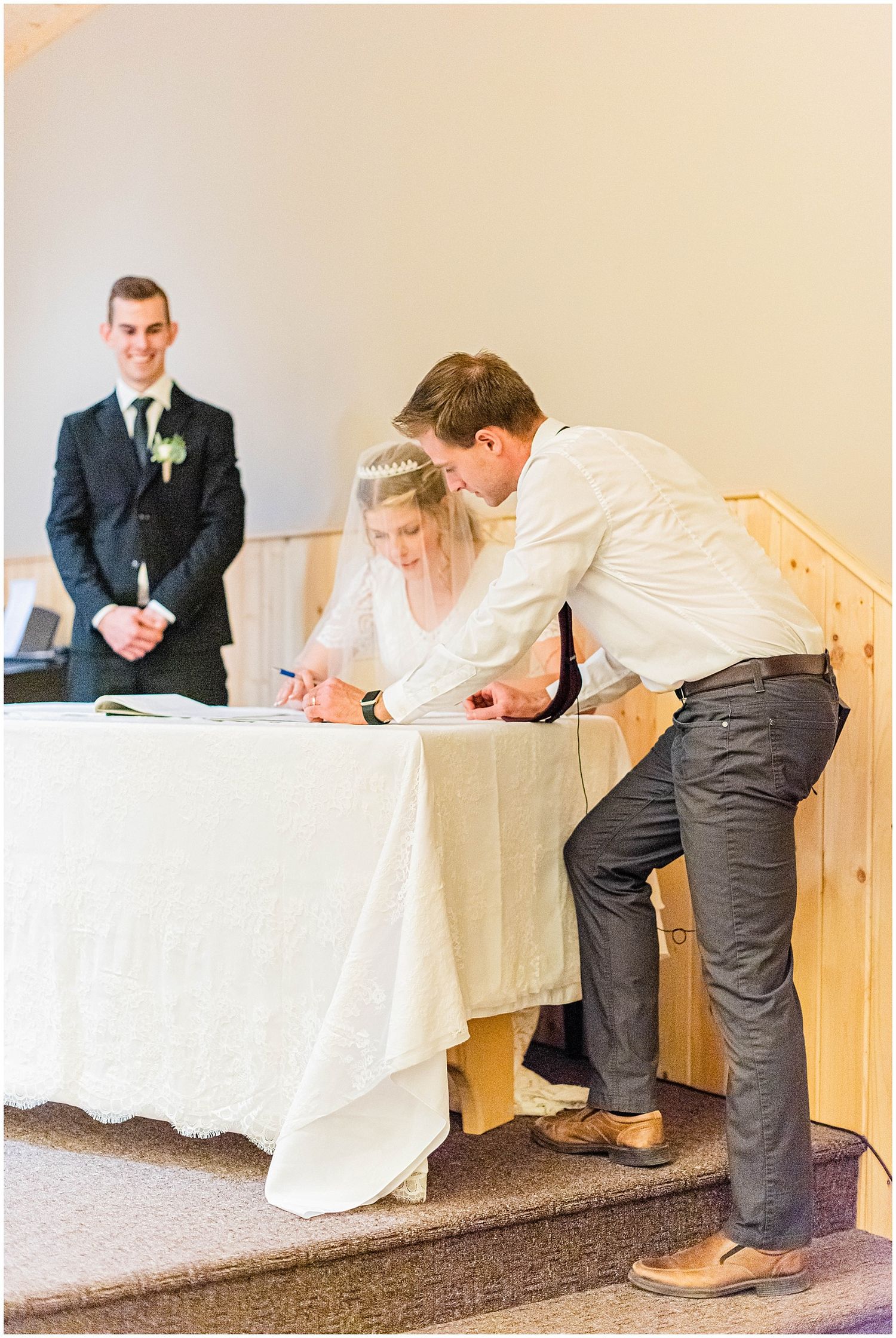 Bride signing wedding registry at white draped table while groom watches during church ceremony.