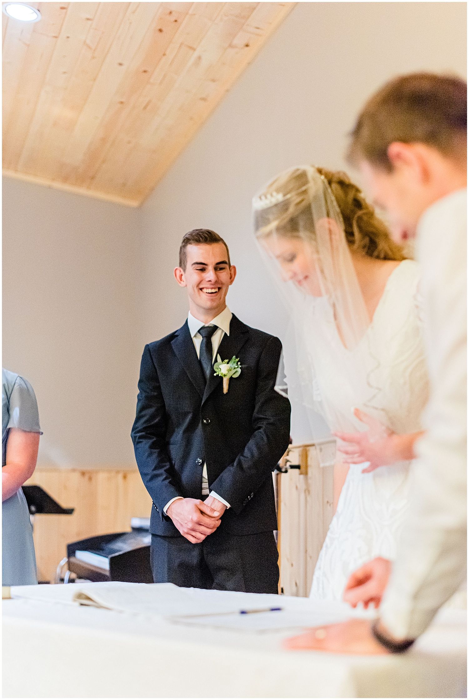 Smiling groom watches bride sign wedding documents during indoor ceremony.